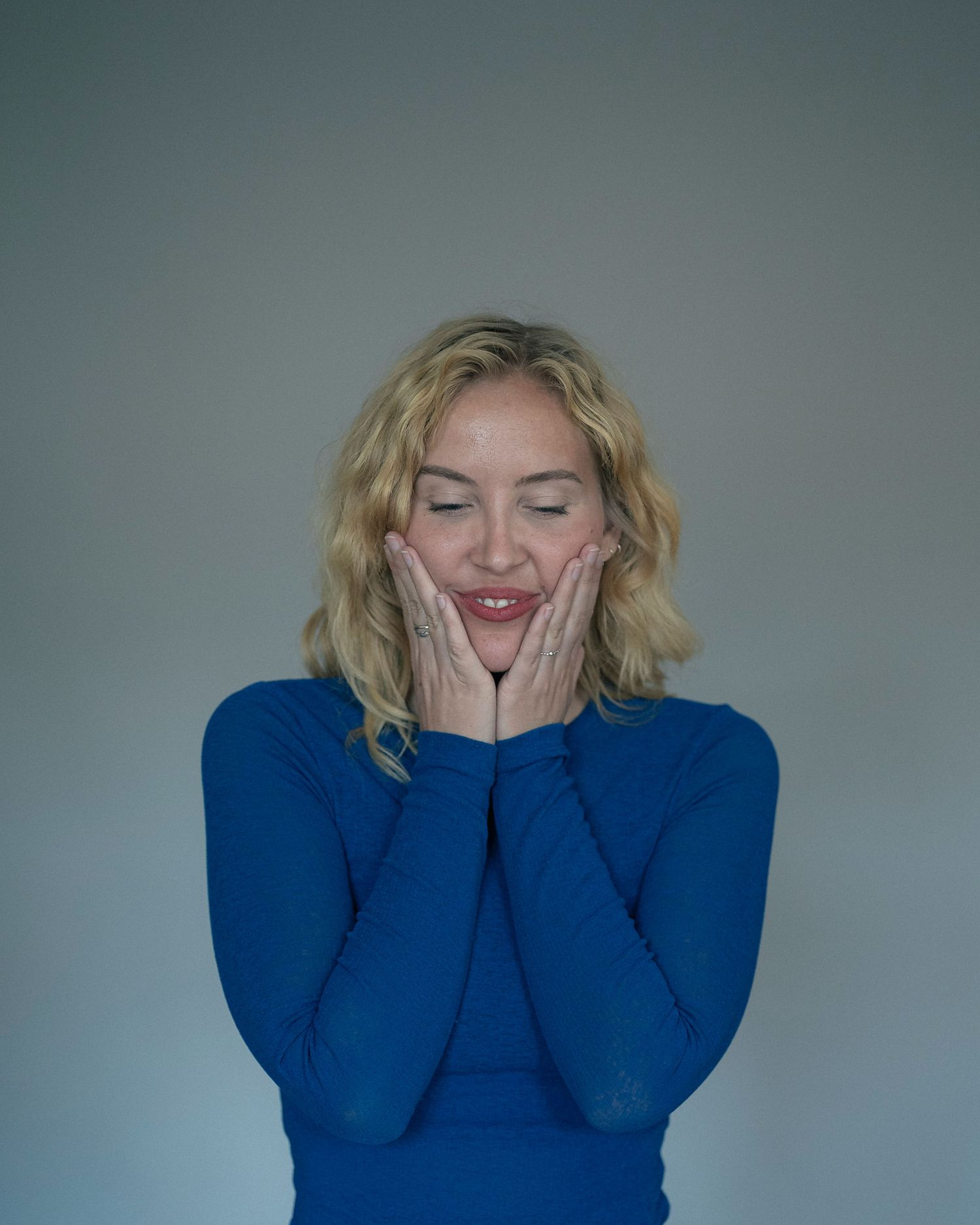 Smiling blonde woman in blue long-sleeve top, hands on cheeks, eyes closed, expressing joy against gray background.