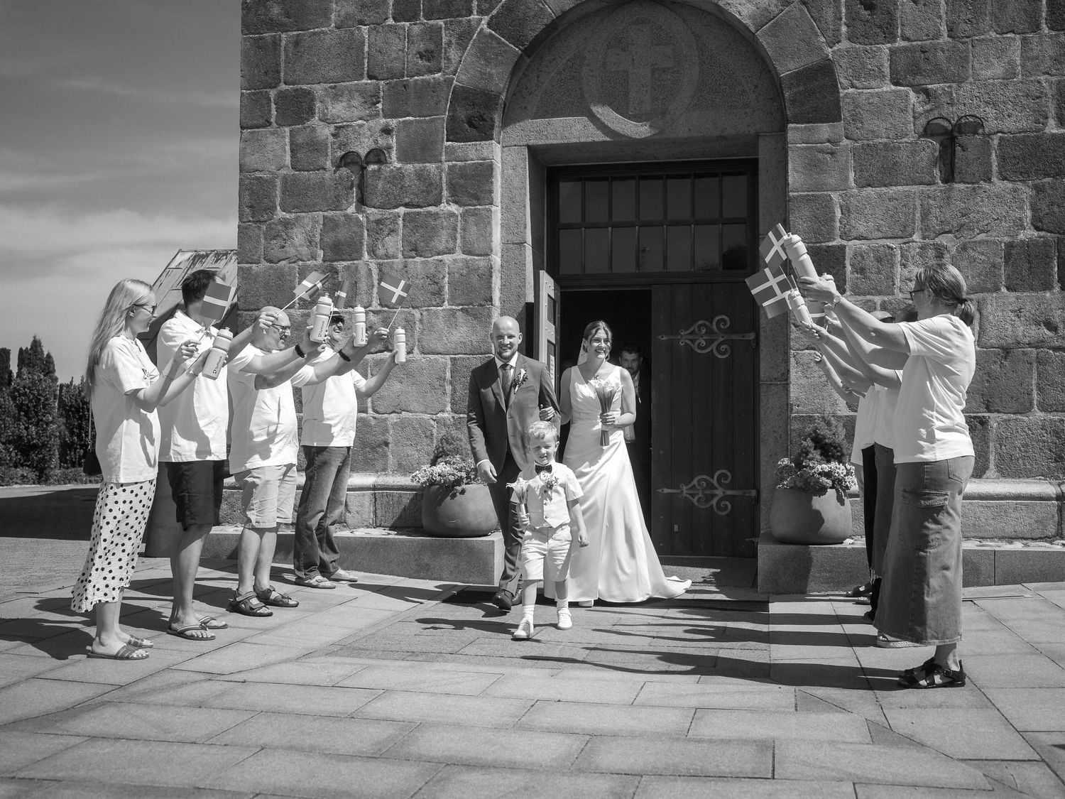 Black and white photo of a wedding couple exiting a stone church, greeted by guests raising glasses in celebration.