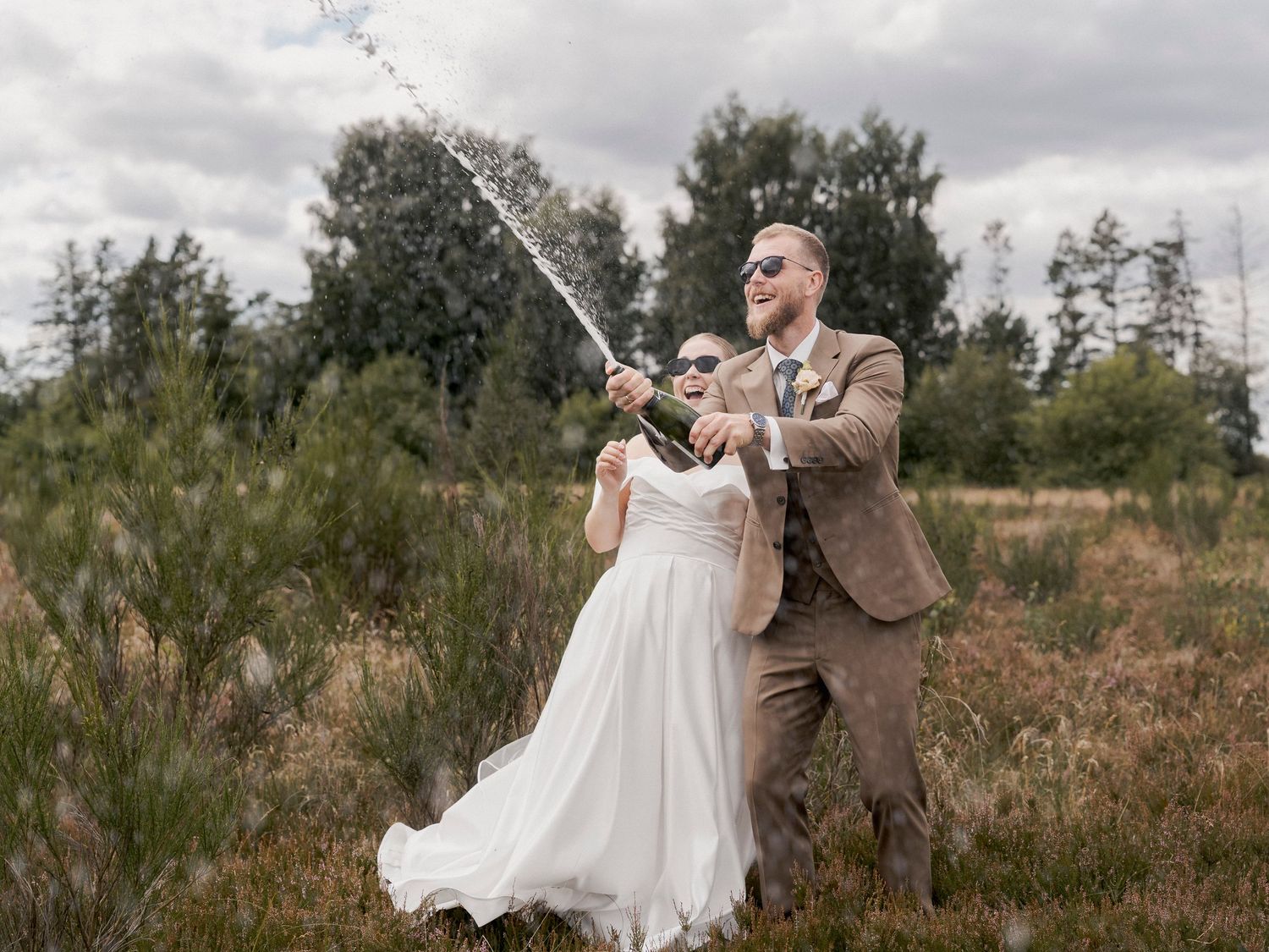 Bride and groom popping champagne bottle in a field, celebrating their wedding day outdoors.
