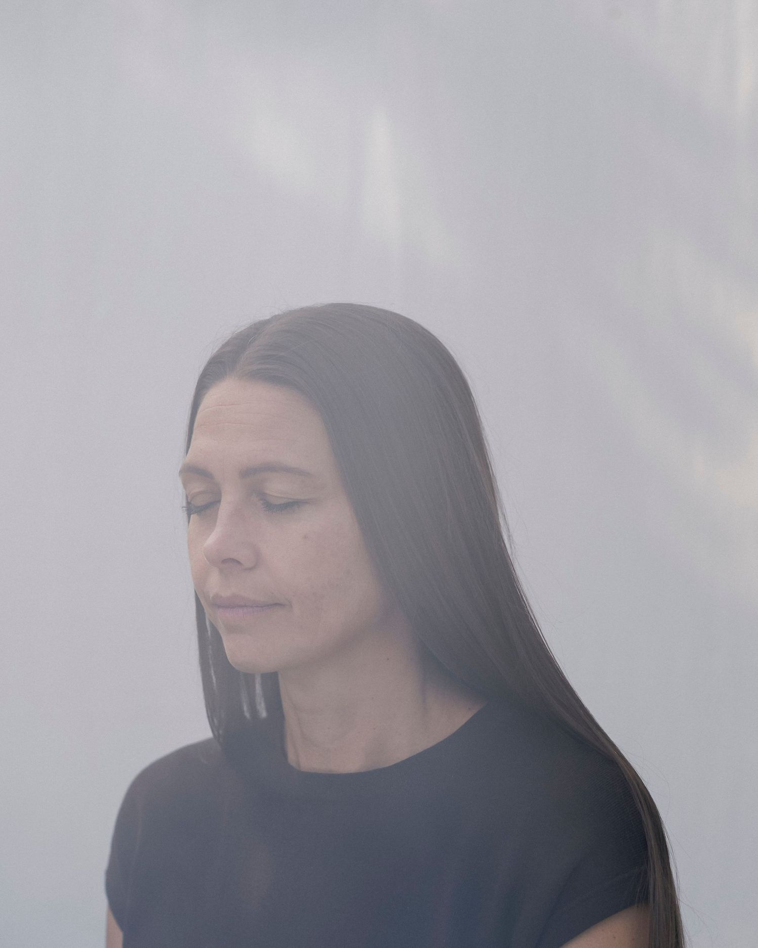 Woman with long dark hair in black top, eyes closed, standing against a soft white background.