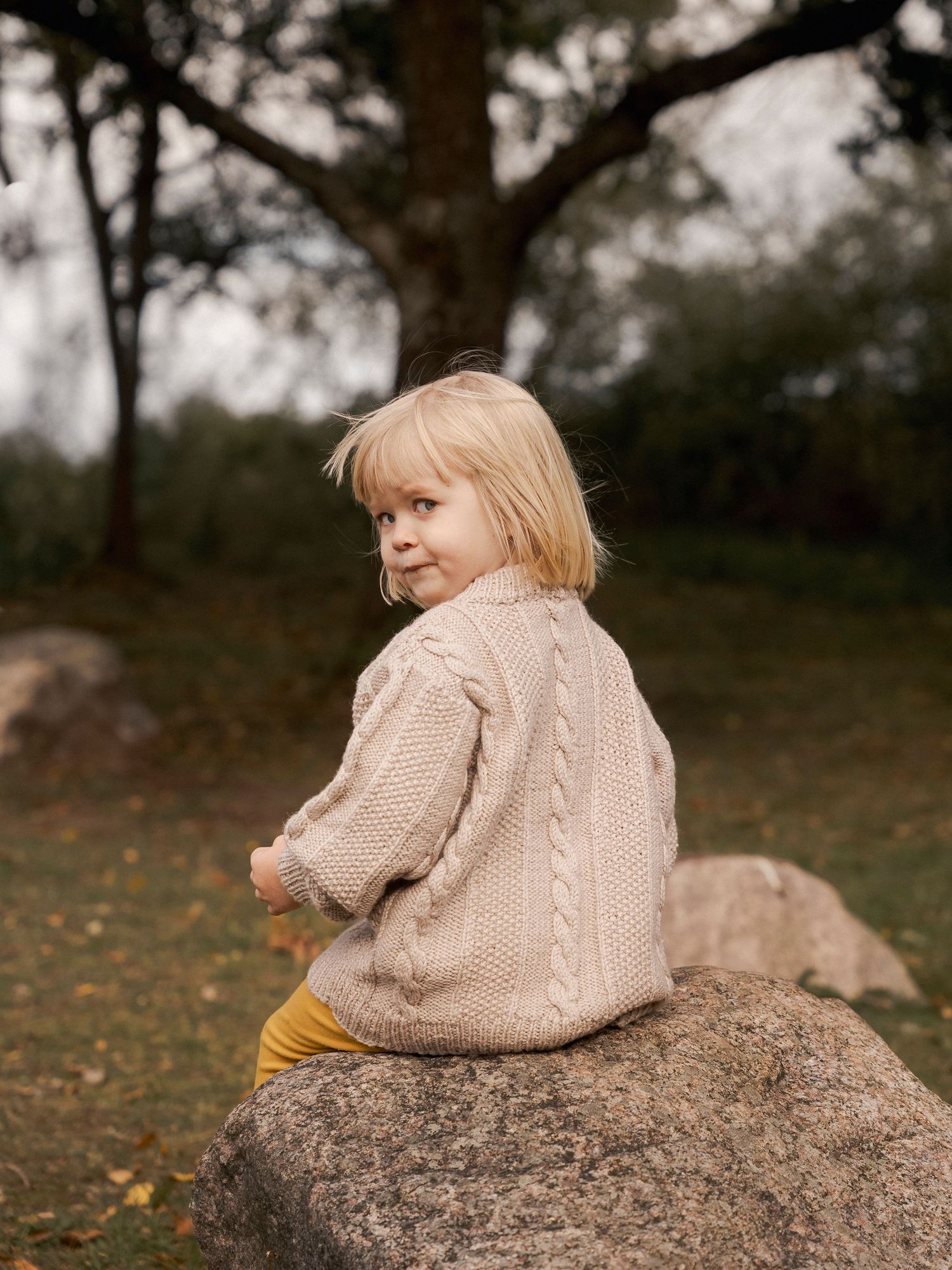 A young blonde child in a beige cable-knit sweater sits on a rock outdoors, looking back over their shoulder.