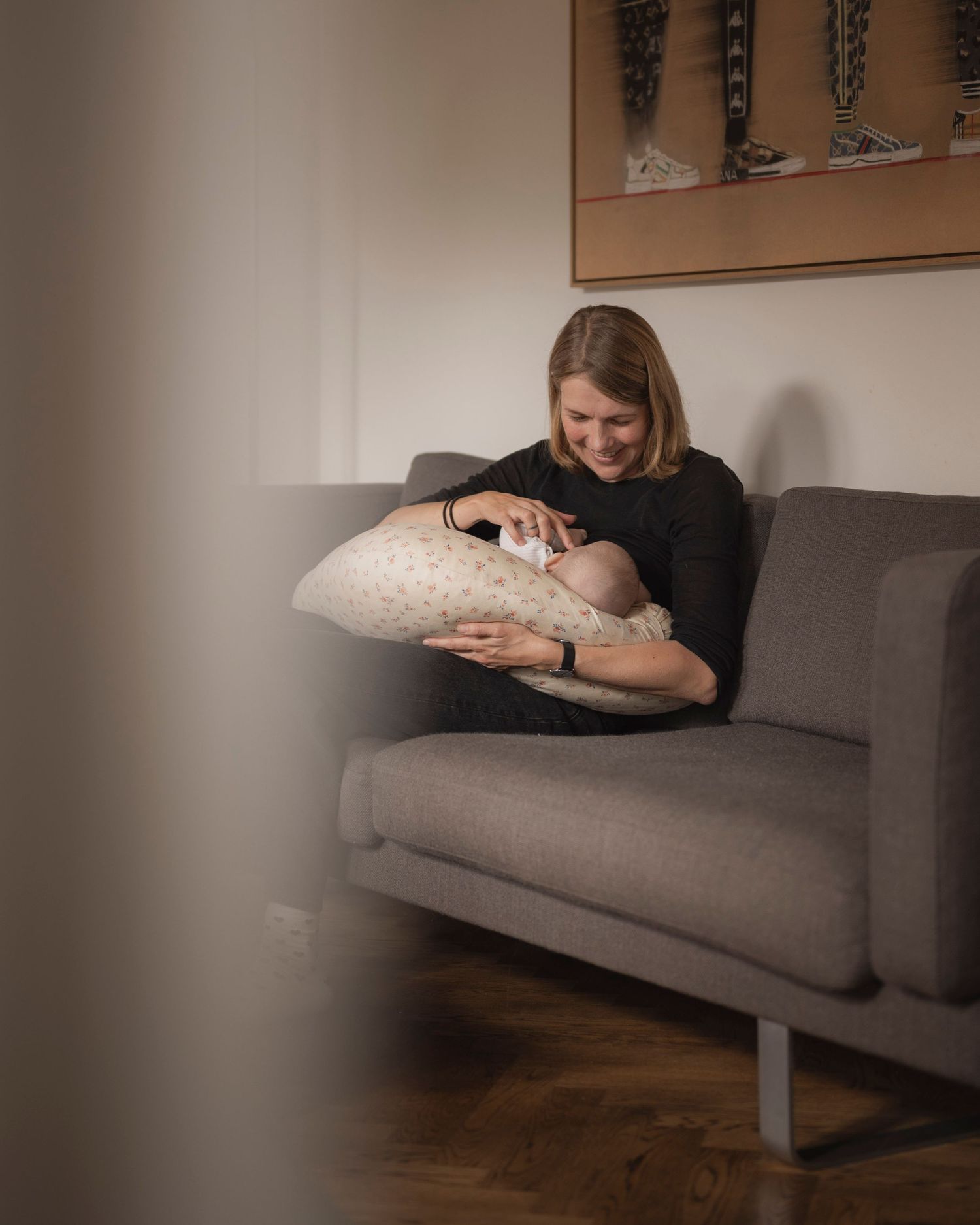 A smiling mother sits on a gray sofa, cradling her newborn baby on a nursing pillow in a cozy home.