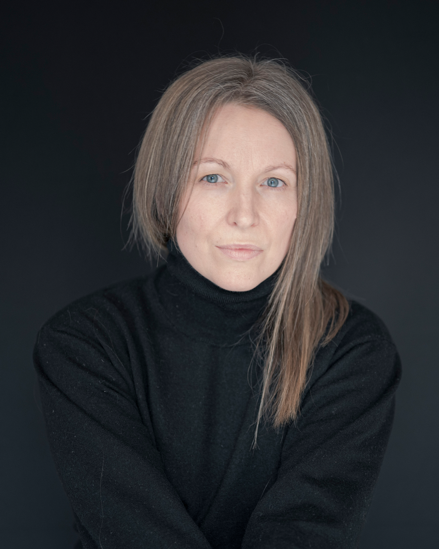 A woman with blonde hair wearing a black turtleneck poses against a dark background.