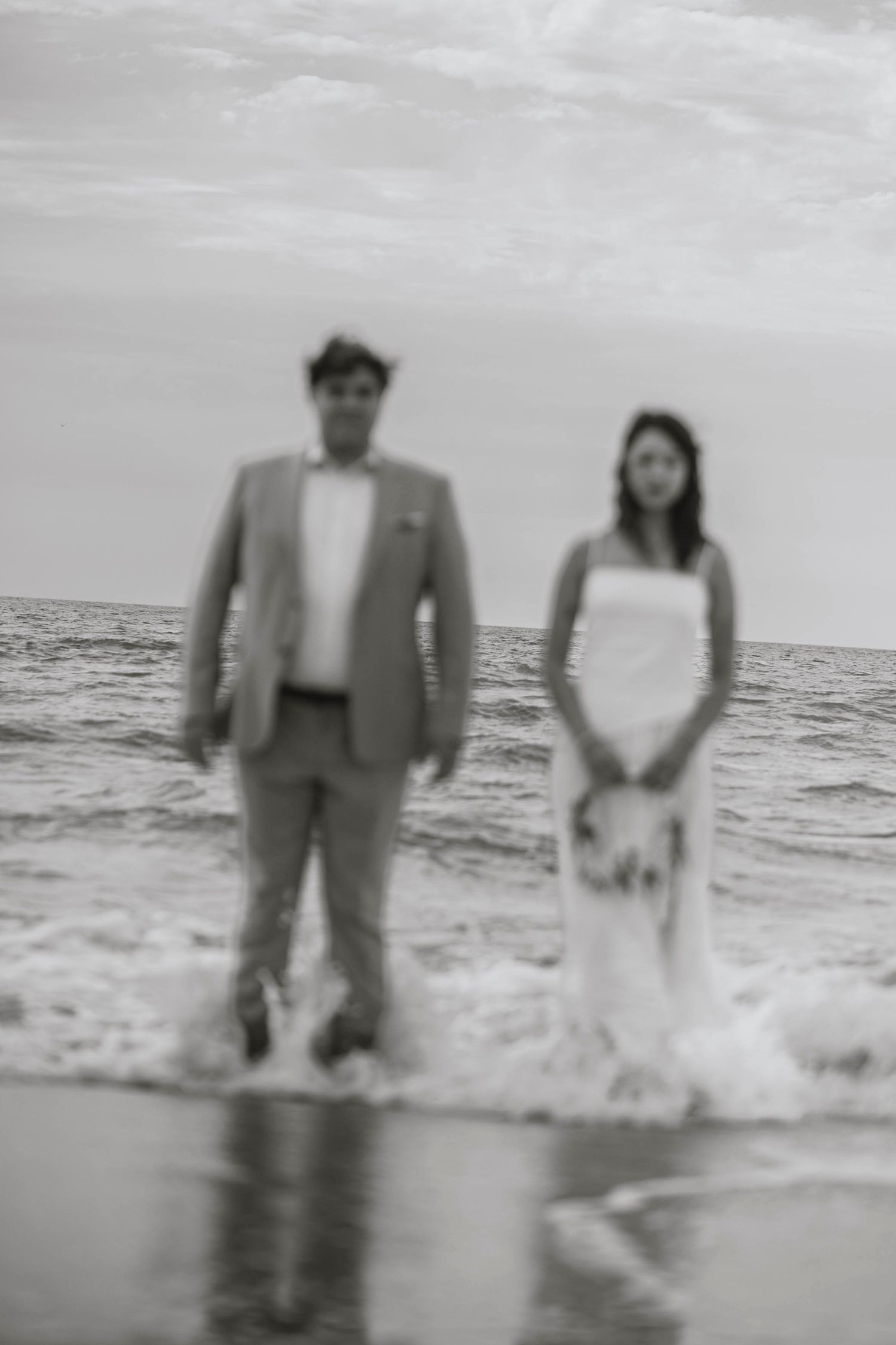 Black and white photo of a bride and groom standing in ocean waves on their wedding day at the beach.
