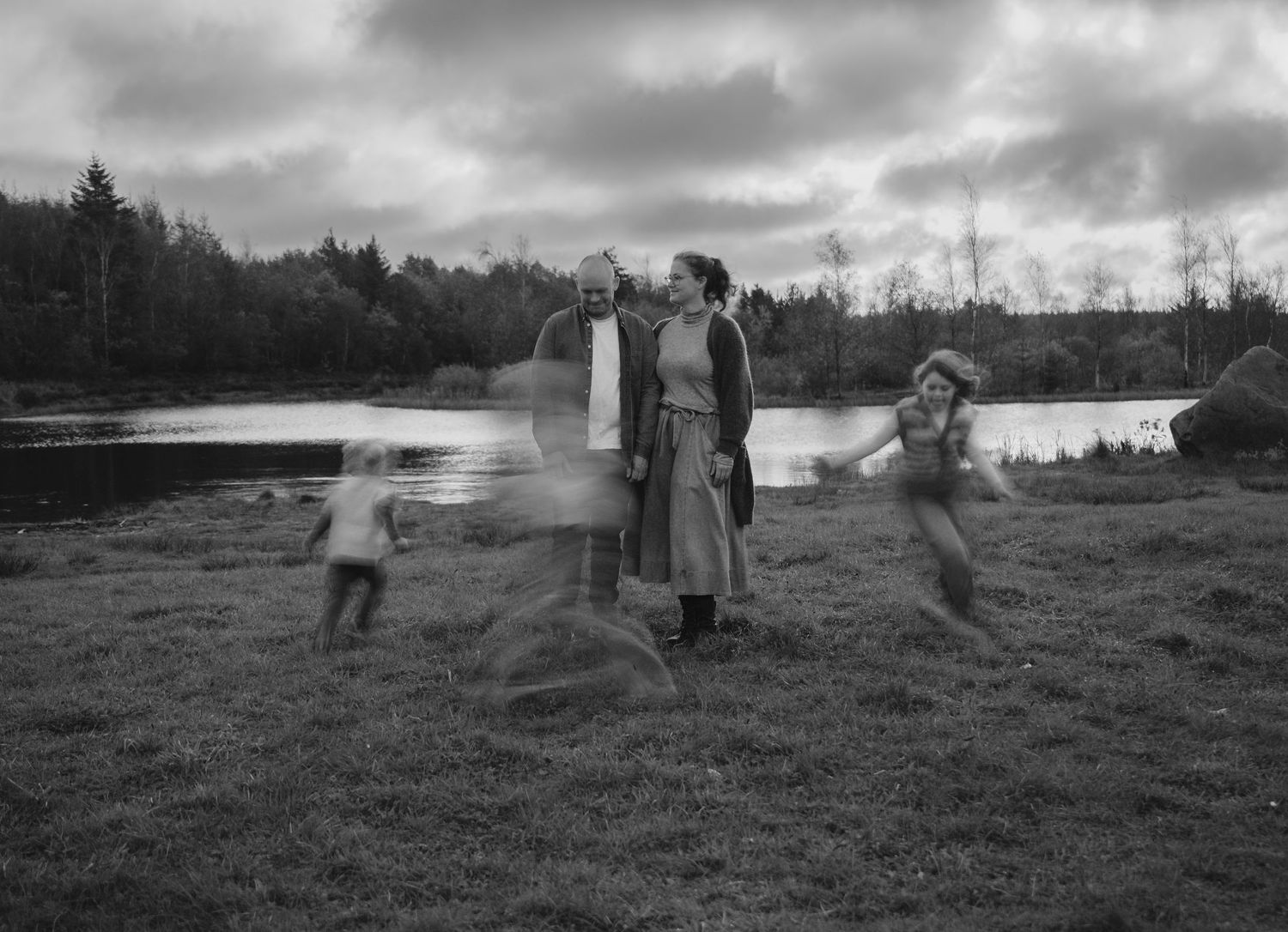 Black and white photo of a family with two children playing outdoors near a pond with trees in background.