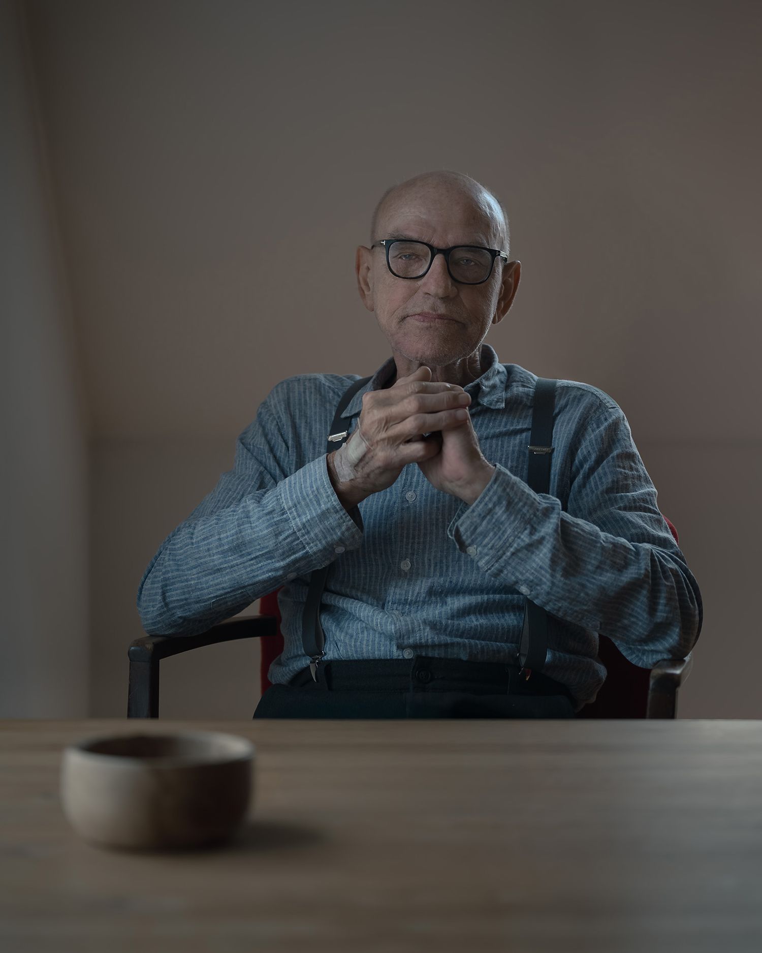 Elderly man with glasses sits at a wooden table with hands clasped, wearing a striped shirt in a dimly lit room.