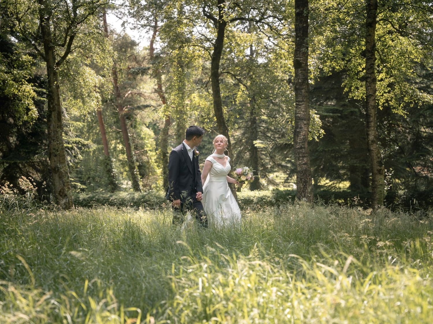 A bride and groom walk hand in hand through a lush green woodland meadow on their wedding day.