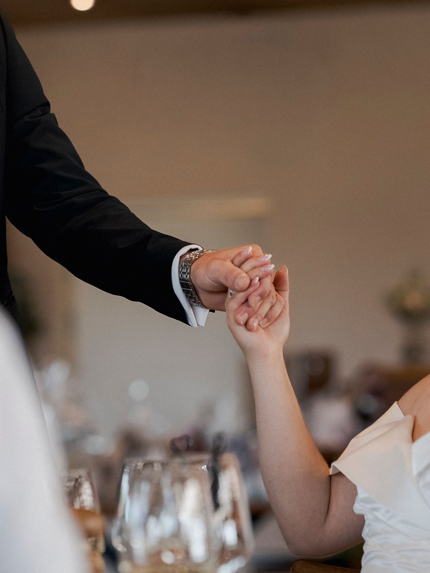A groom in a black suit holds hands with a bride in a white dress at a wedding reception.