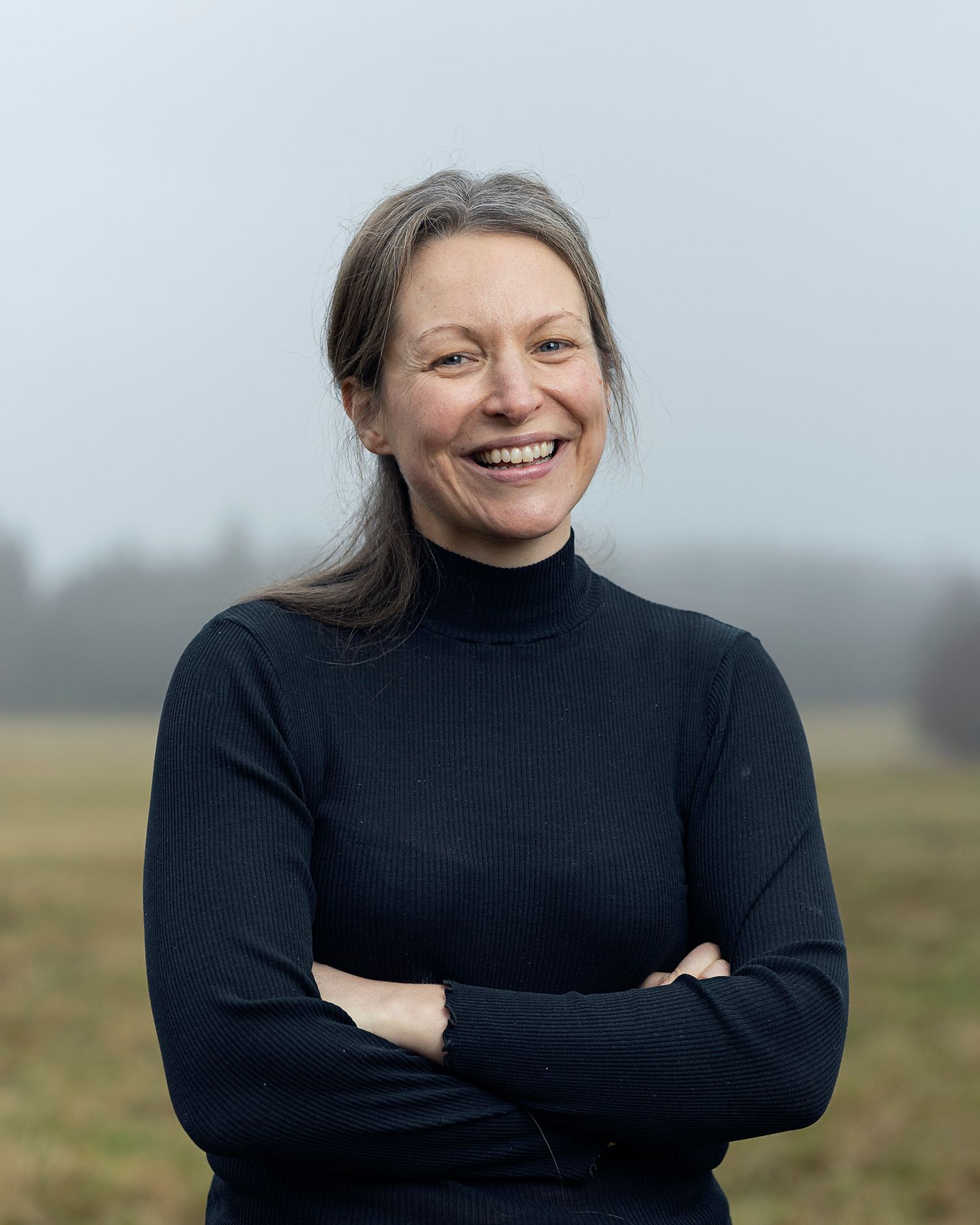 Smiling woman with gray hair in black turtleneck sweater standing outdoors with arms crossed on a foggy day.