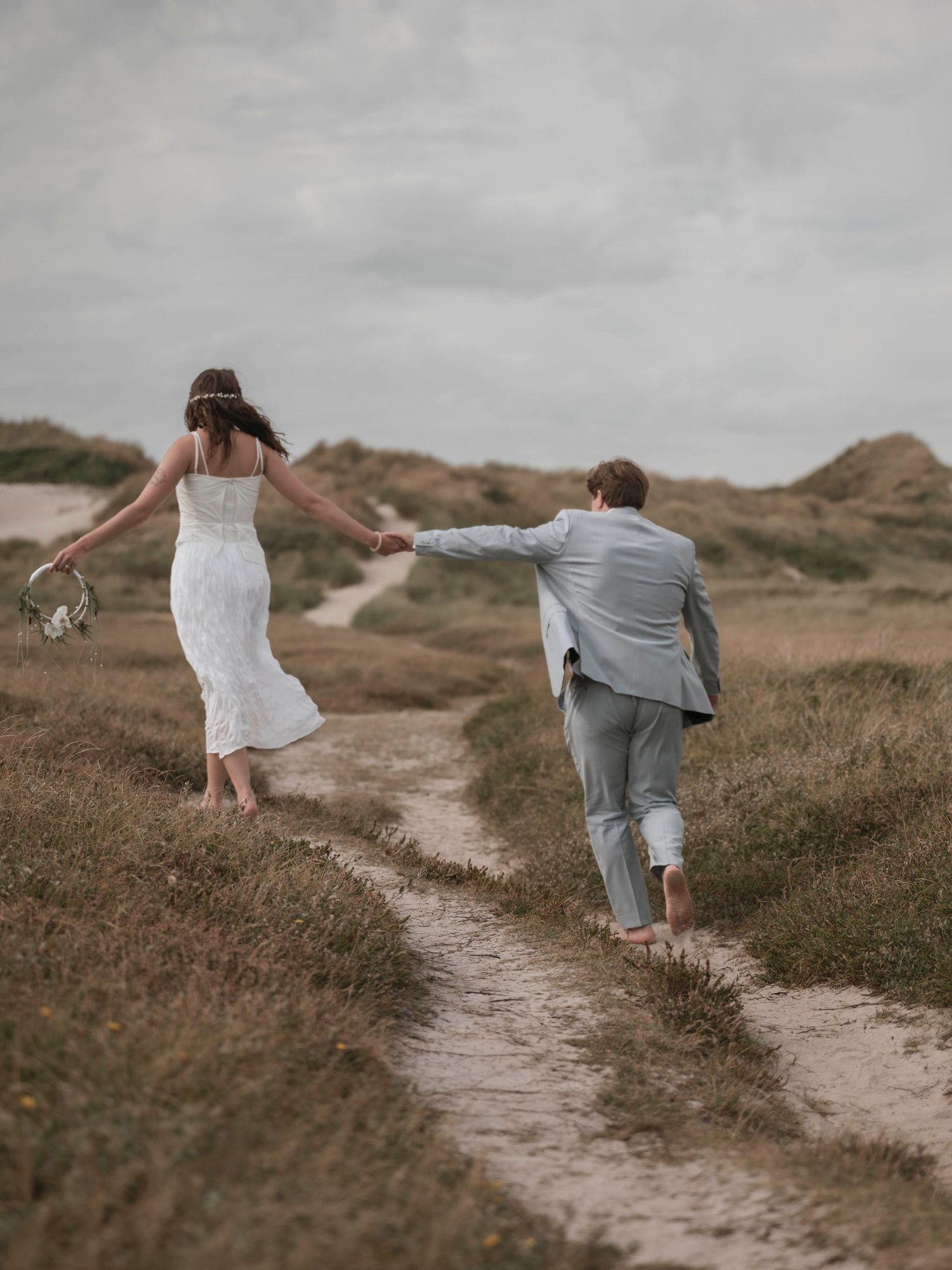 Barefoot bride and groom holding hands walking along sandy dune path on overcast day.