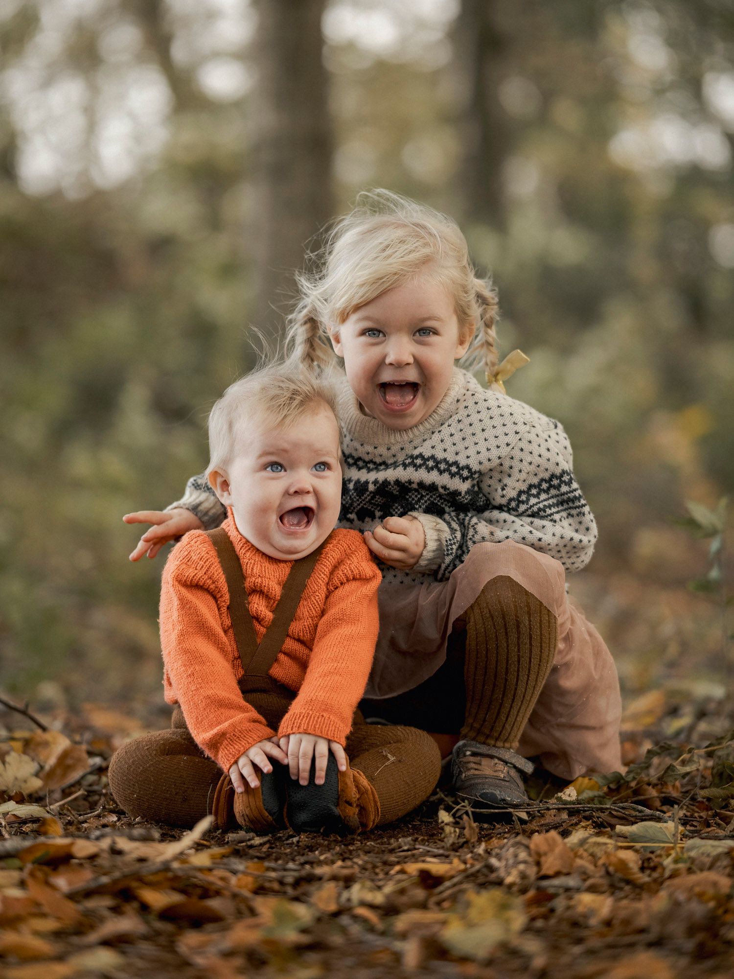 Two laughing toddlers sit together on autumn leaves in a forest, wearing cozy knit sweaters in orange and beige.