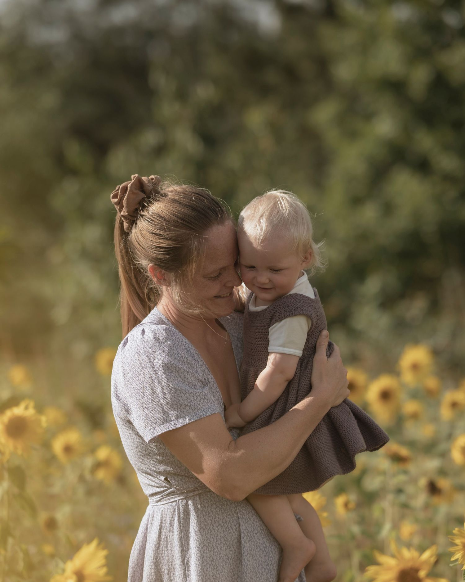 A mother tenderly holds her toddler in a sunny sunflower field, both sharing a sweet moment outdoors.
