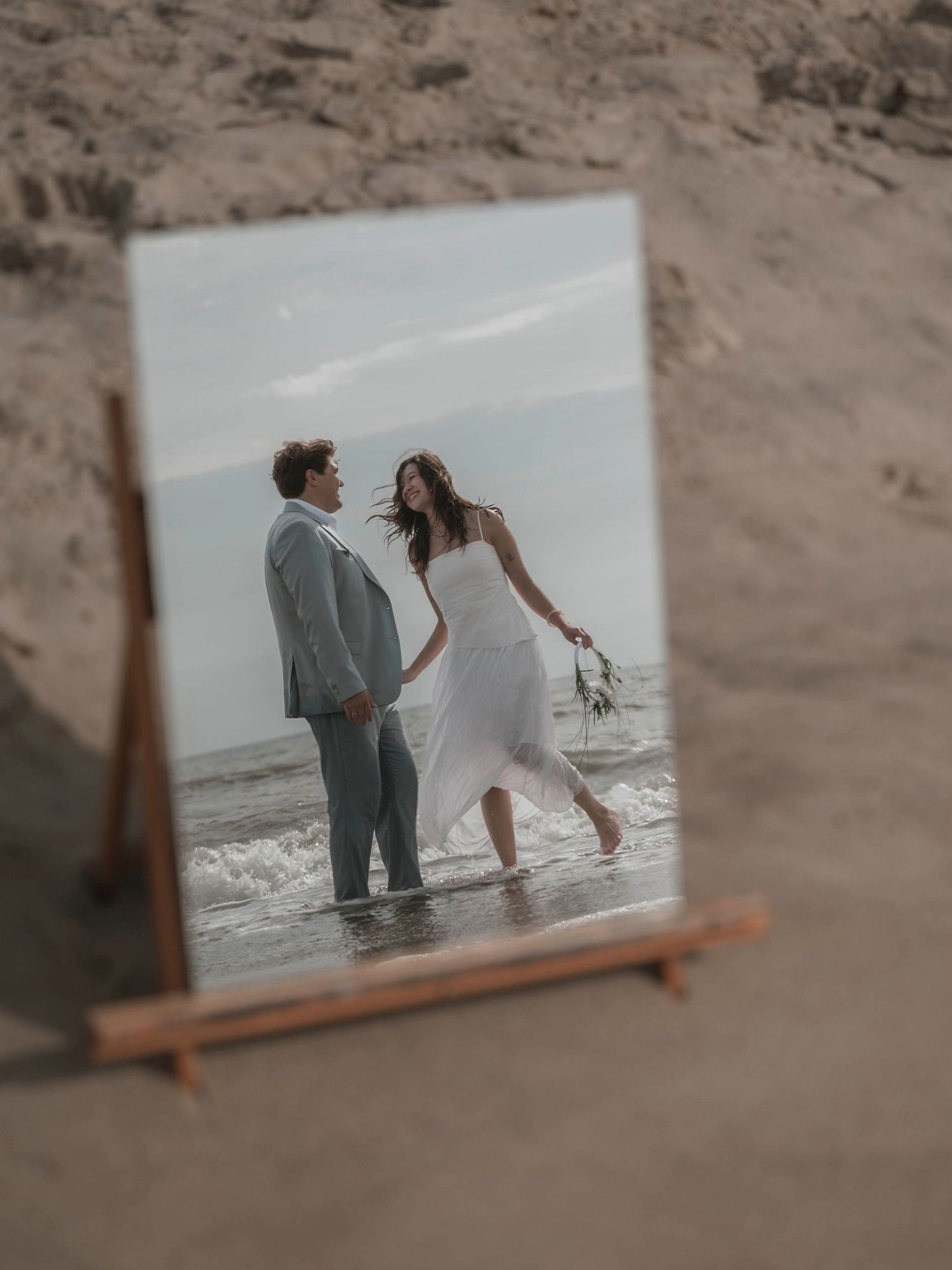 A wedding couple holds hands on a beach shoreline, reflected in a mirror propped on sand.