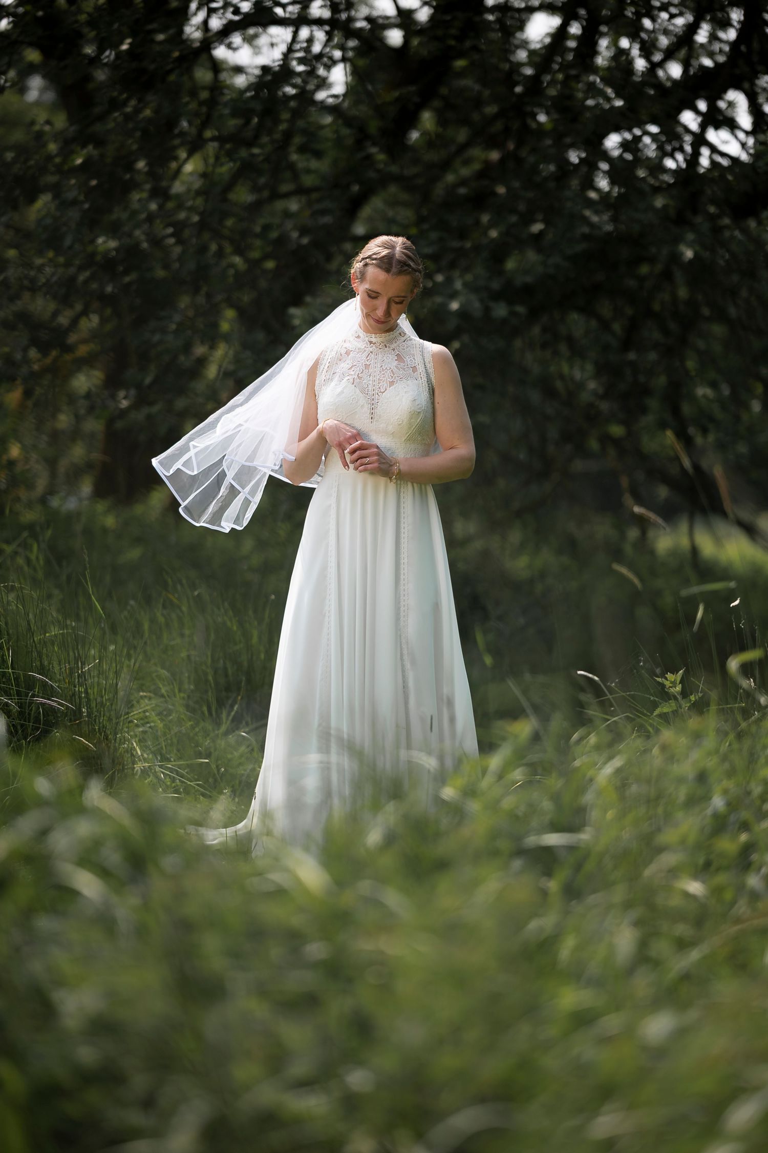 Bride in white lace gown and veil stands gracefully among lush green trees and tall grass in an outdoor setting.