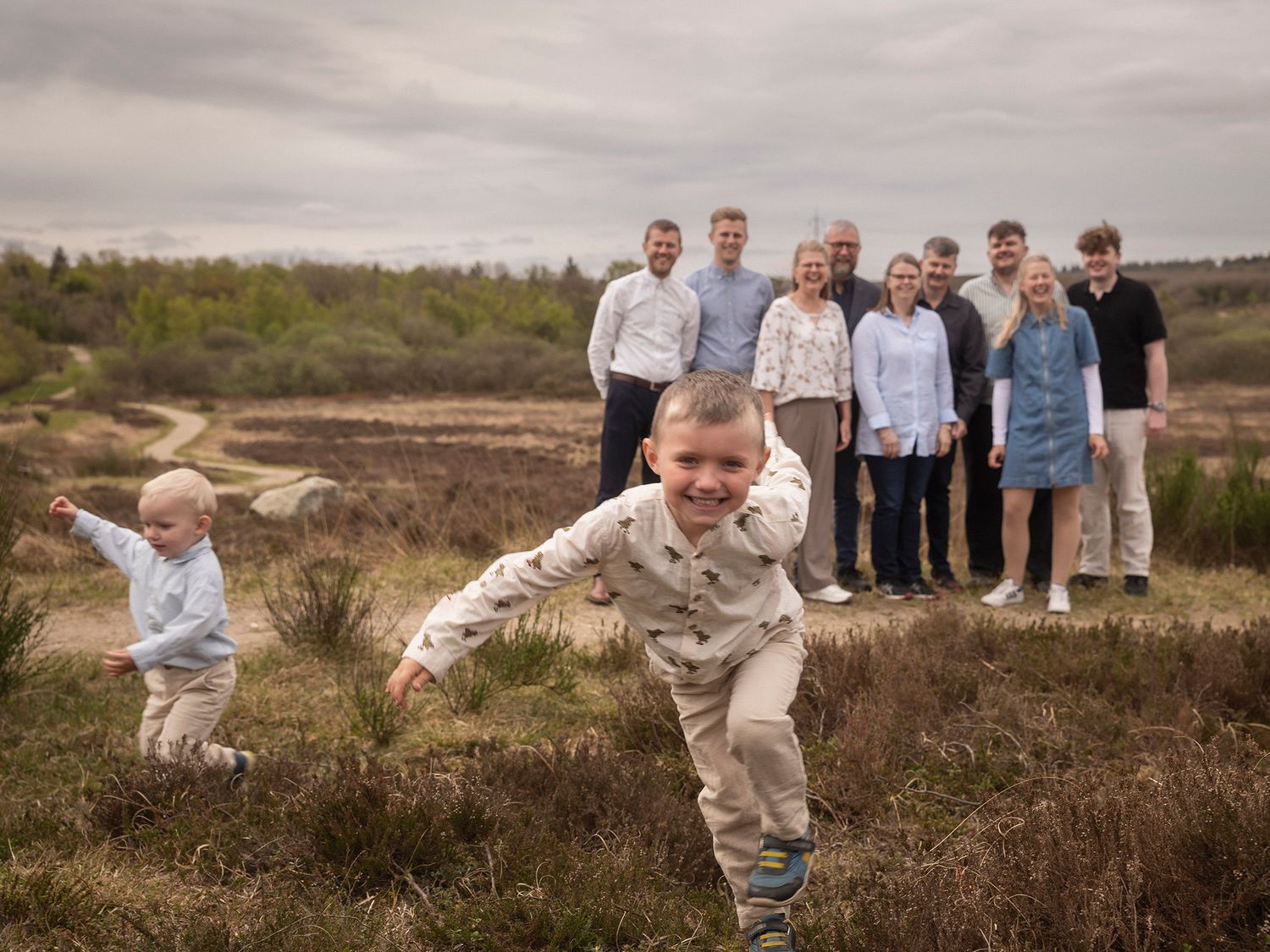 Two young boys run joyfully through moorland while a large family group poses together in the background.