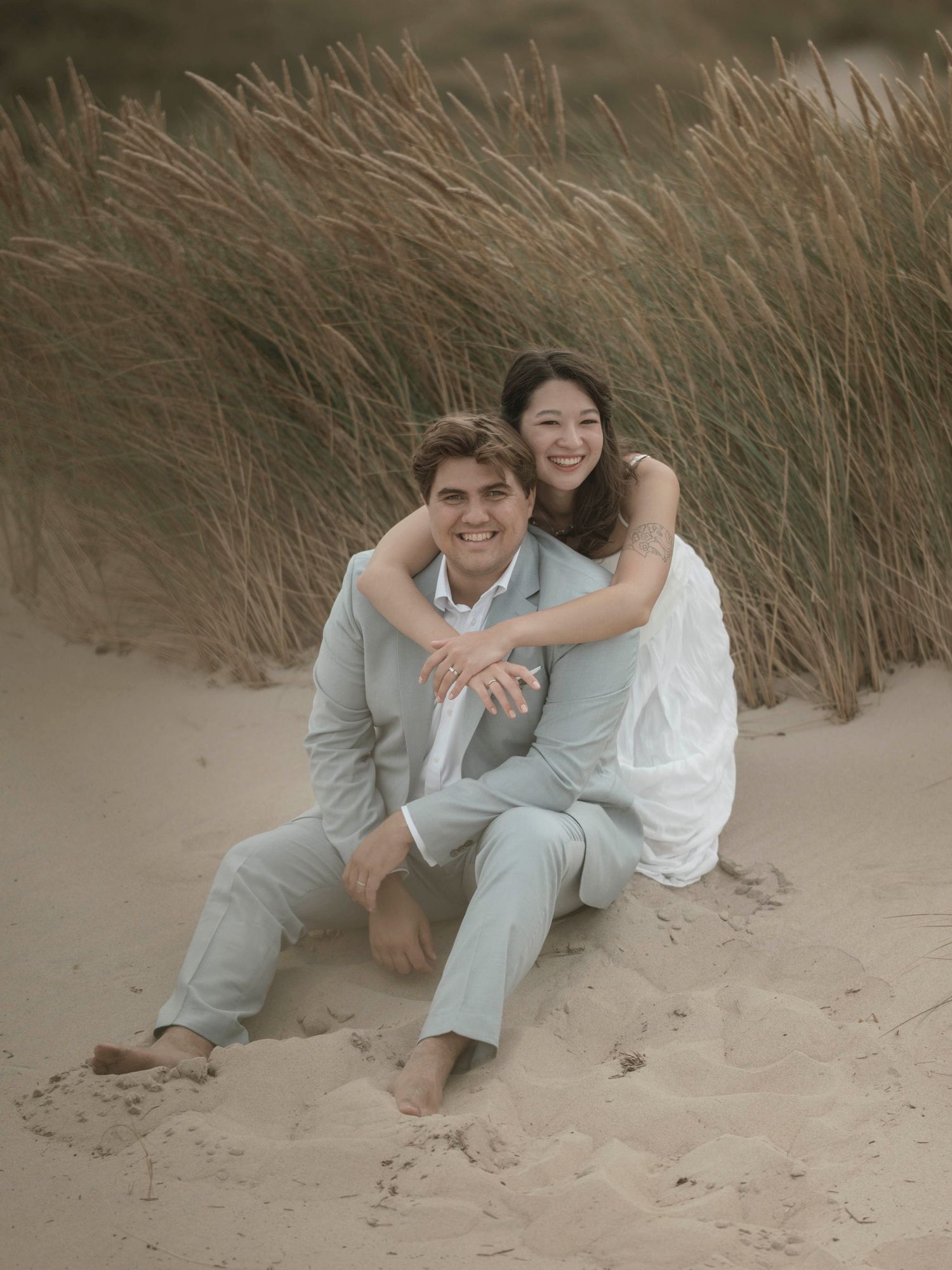 Couple relaxing on sandy beach among coastal grass during wedding photoshoot.