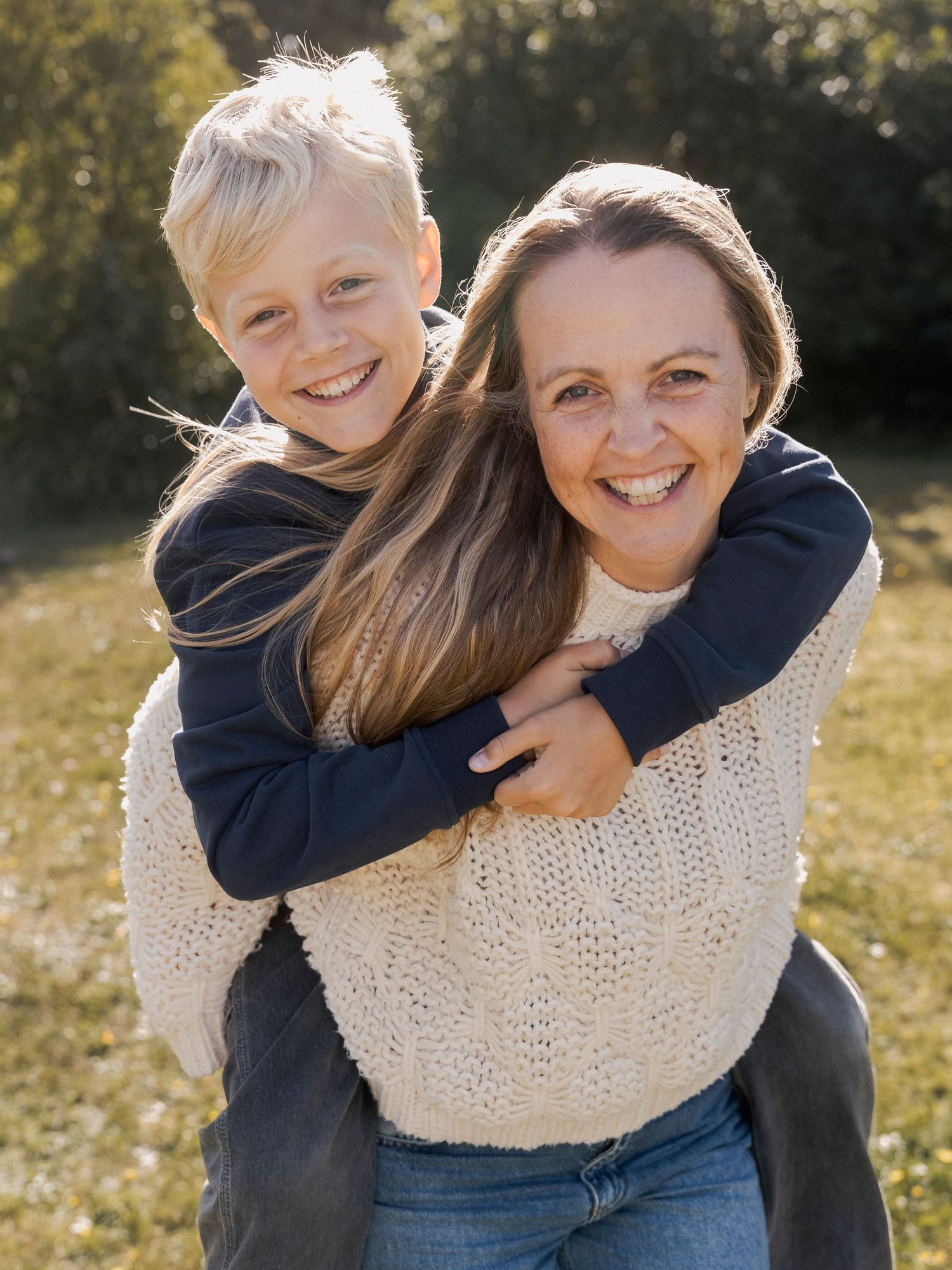 A woman gives a smiling blonde boy a piggyback ride outdoors in a sunny green field, both laughing joyfully.