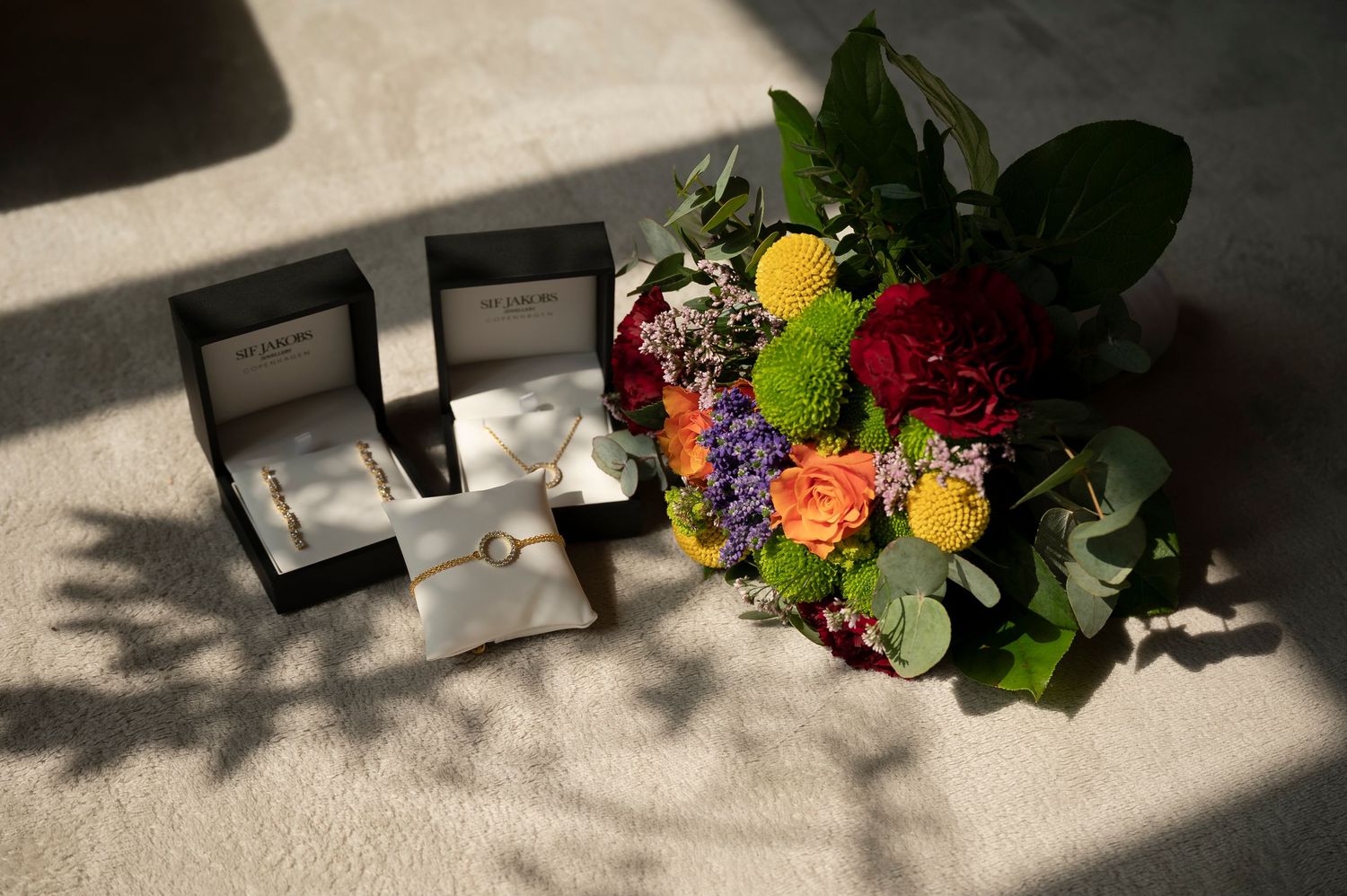 Wedding rings in open boxes beside a colorful bridal bouquet with red, orange, and yellow flowers on a white surface.