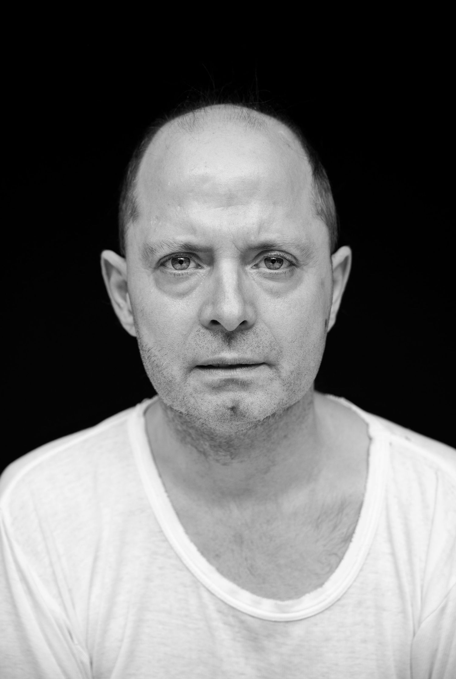 Black and white portrait of a bald middle-aged man in a white t-shirt against a dark background.