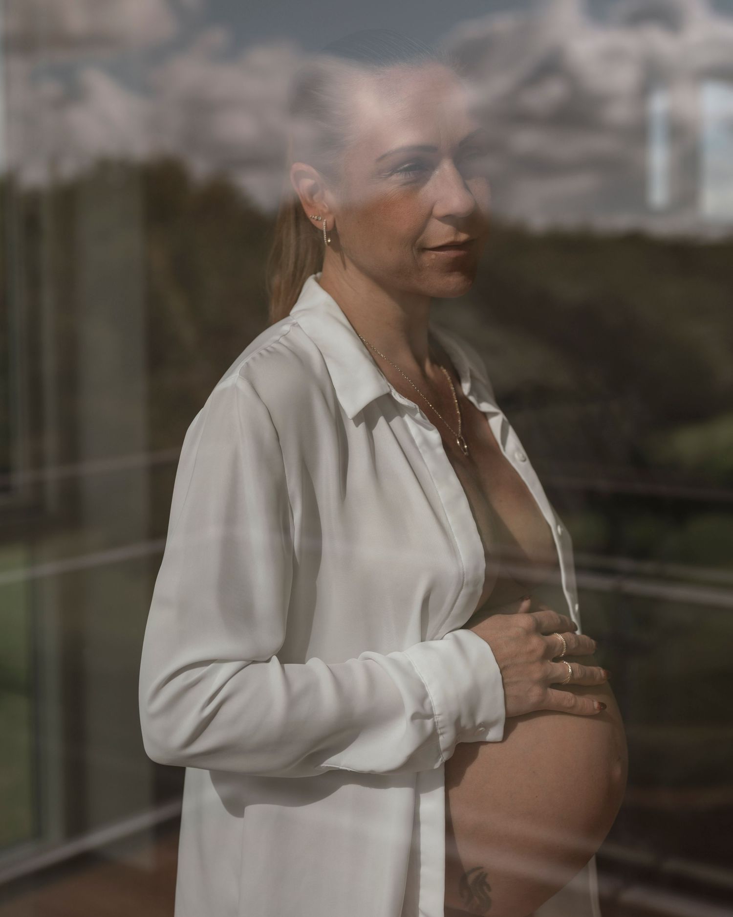 Pregnant woman in white shirt cradling baby bump, standing by glass window with green landscape reflection.