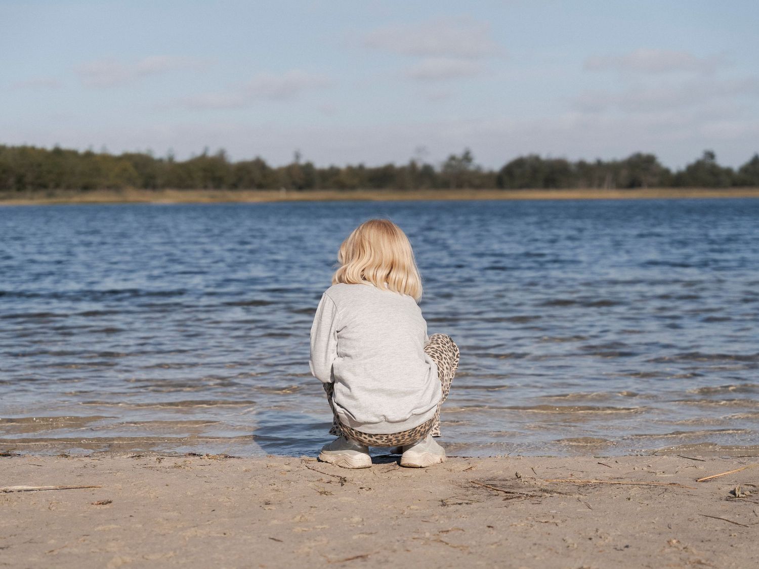 A young blonde child crouching alone on a sandy beach shore, gazing at calm blue water under a partly cloudy sky.