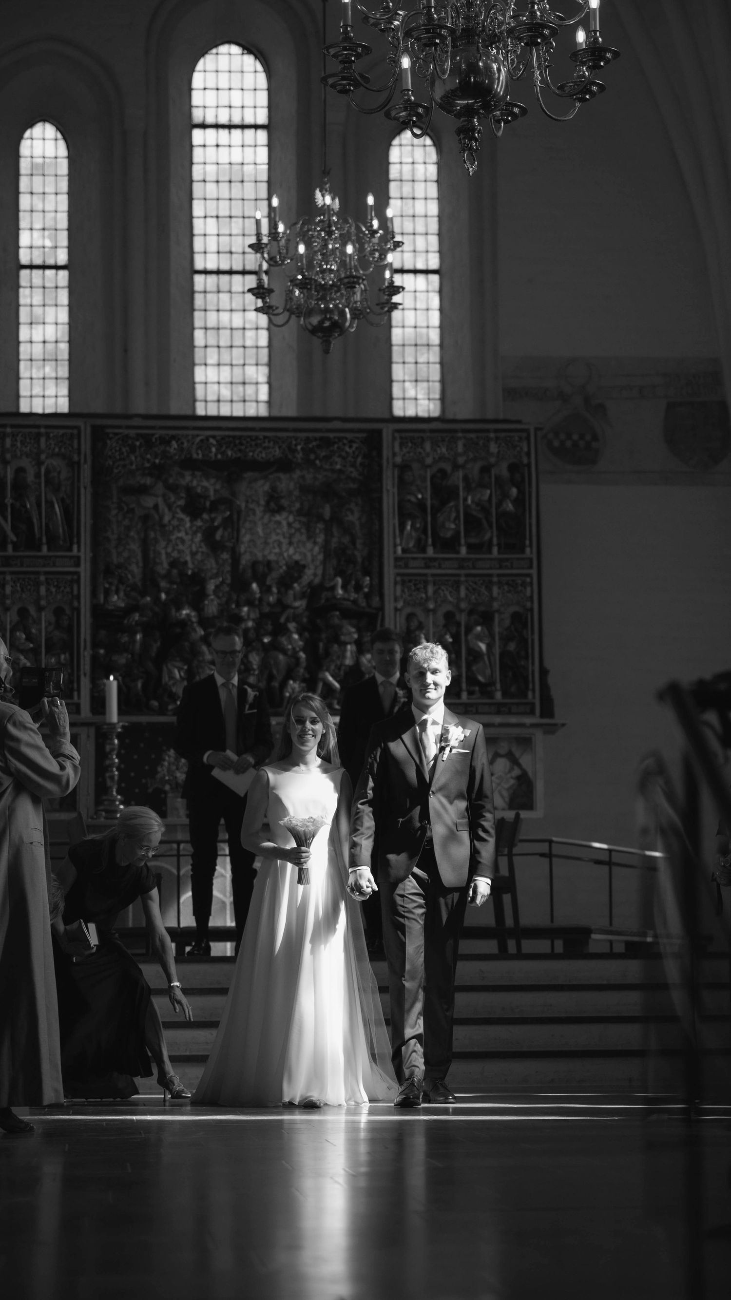 Black and white photo of bride and groom walking down church aisle under chandelier during wedding ceremony.