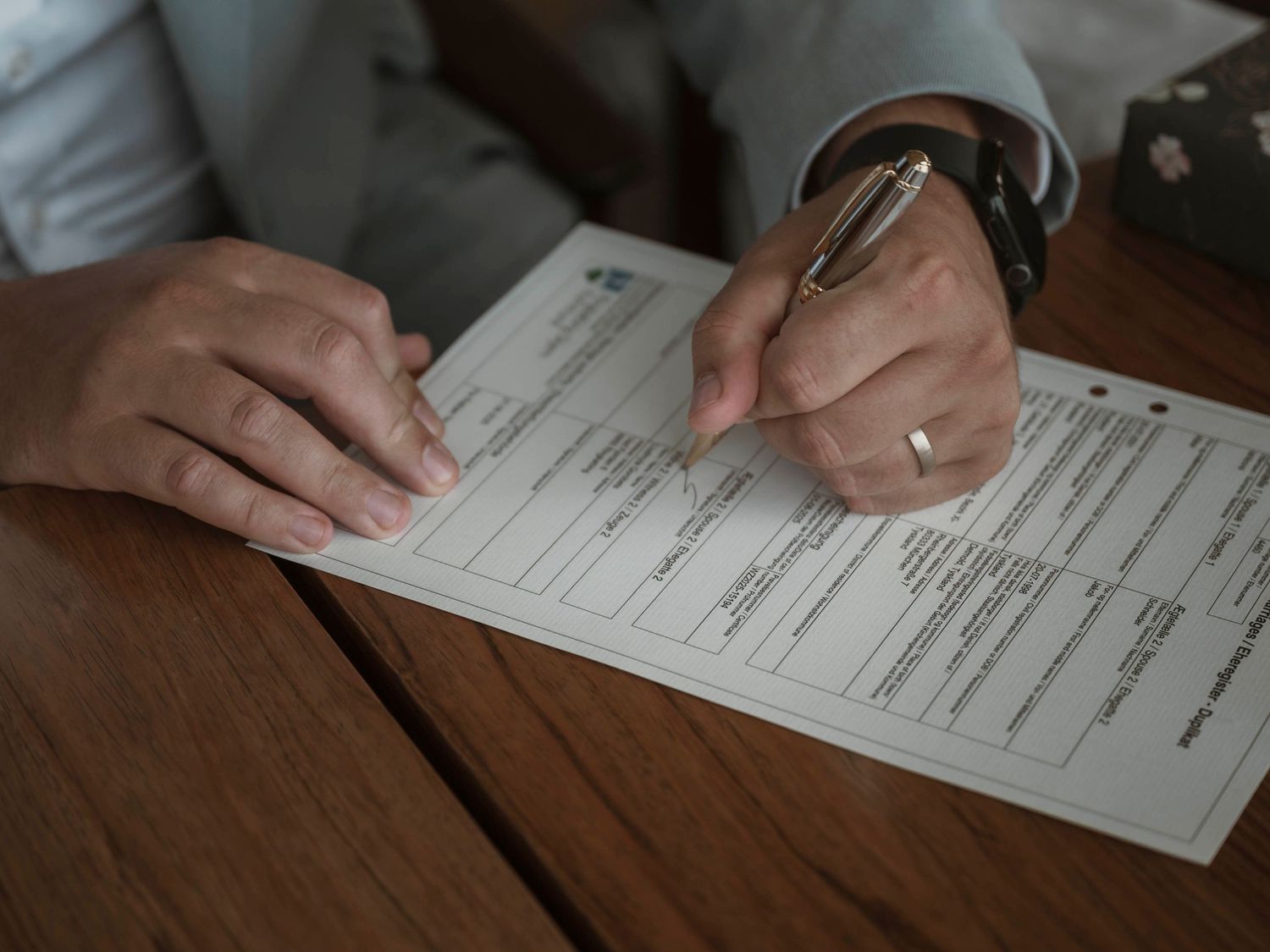 Hands holding pens over paperwork on a wooden table surface in dim lighting.