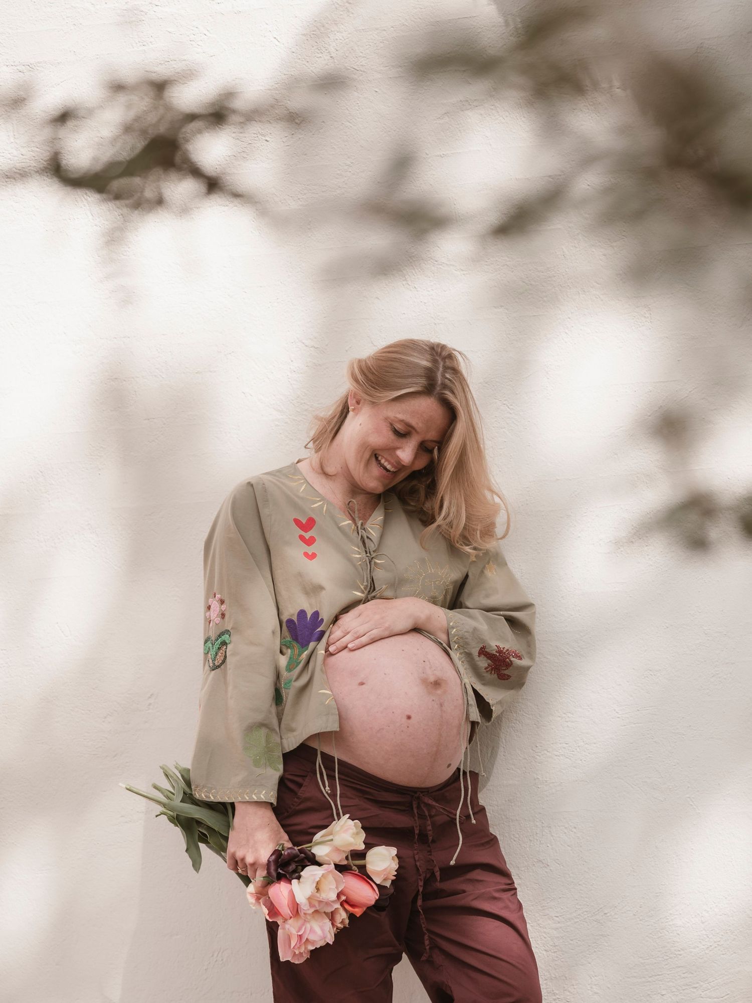 Pregnant woman in floral top holds flowers, smiling down at baby bump against white wall.