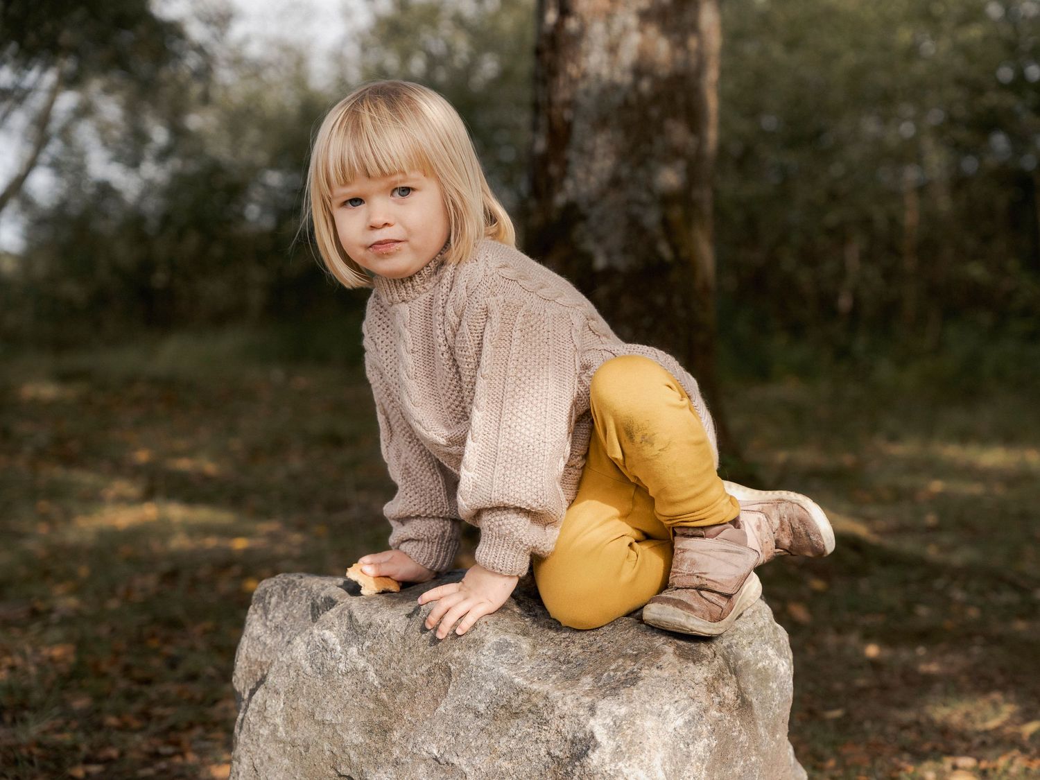 Young blonde child in beige sweater and mustard pants sitting on a large rock outdoors in a forest setting.