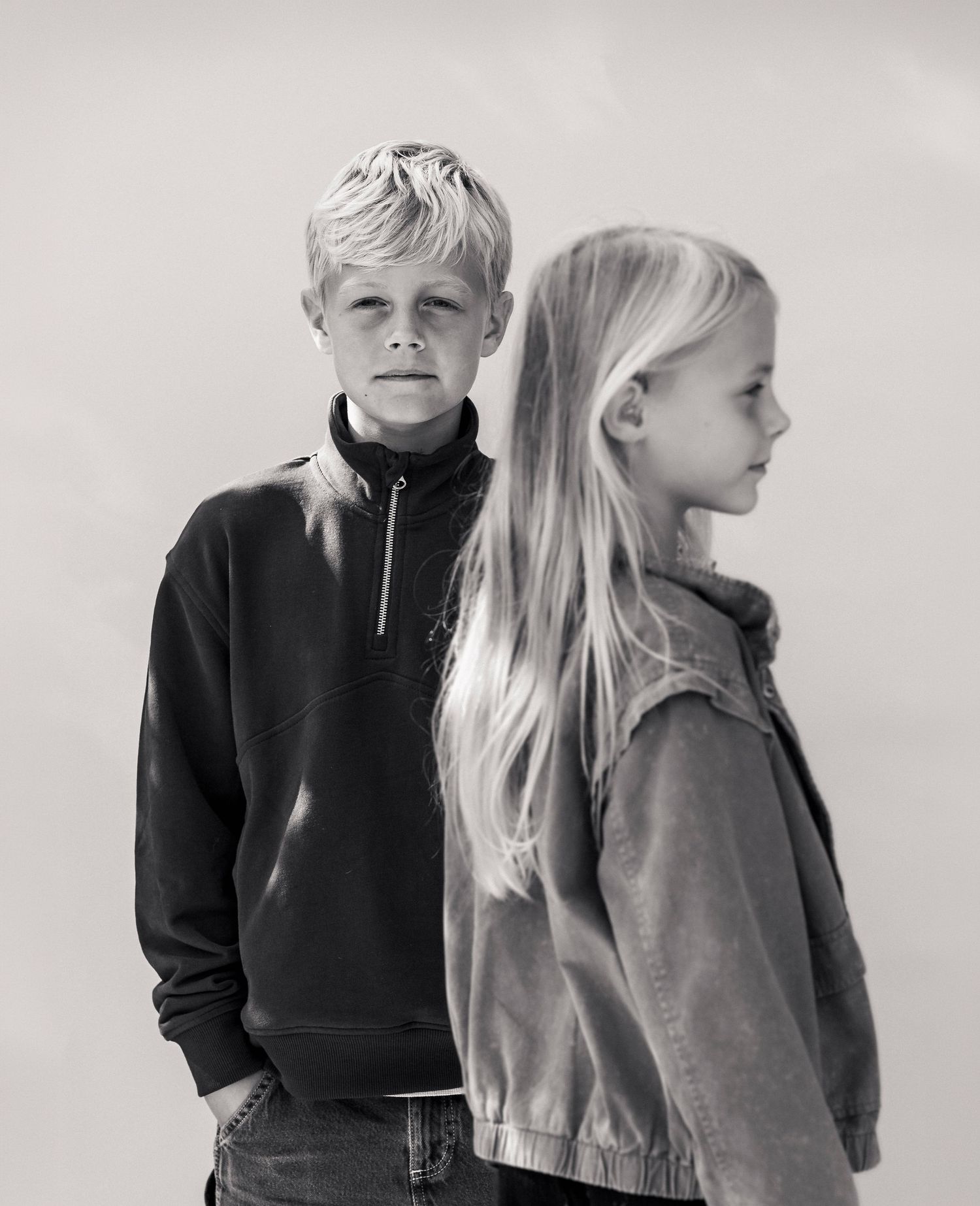 Black and white portrait of two blonde children, a boy in a sweatshirt and a girl in a jacket, posing outdoors.