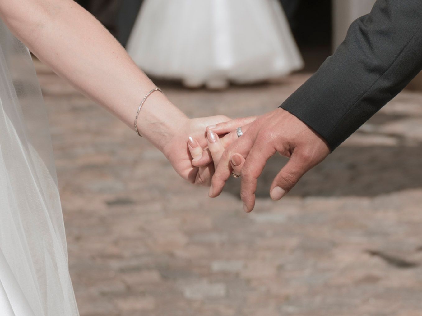 Bride and groom holding hands, showcasing wedding rings and bridal veil at an outdoor wedding ceremony.