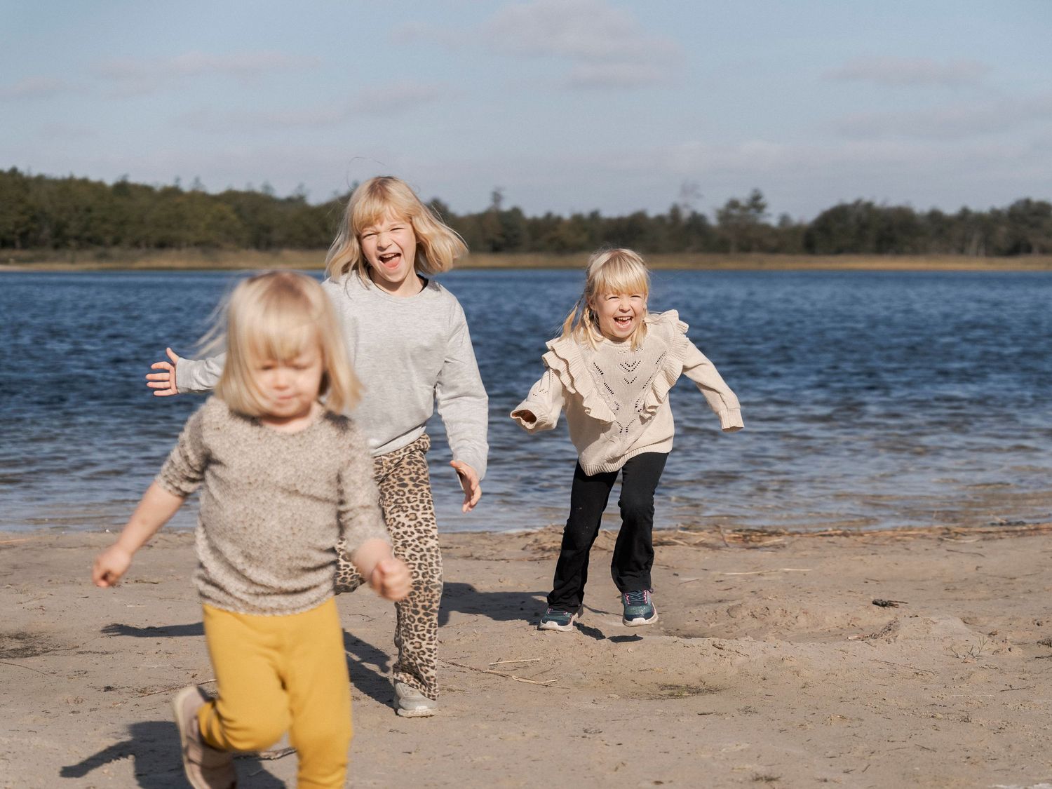 Three young children laughing and running on a sandy beach beside a calm lake on a sunny day.
