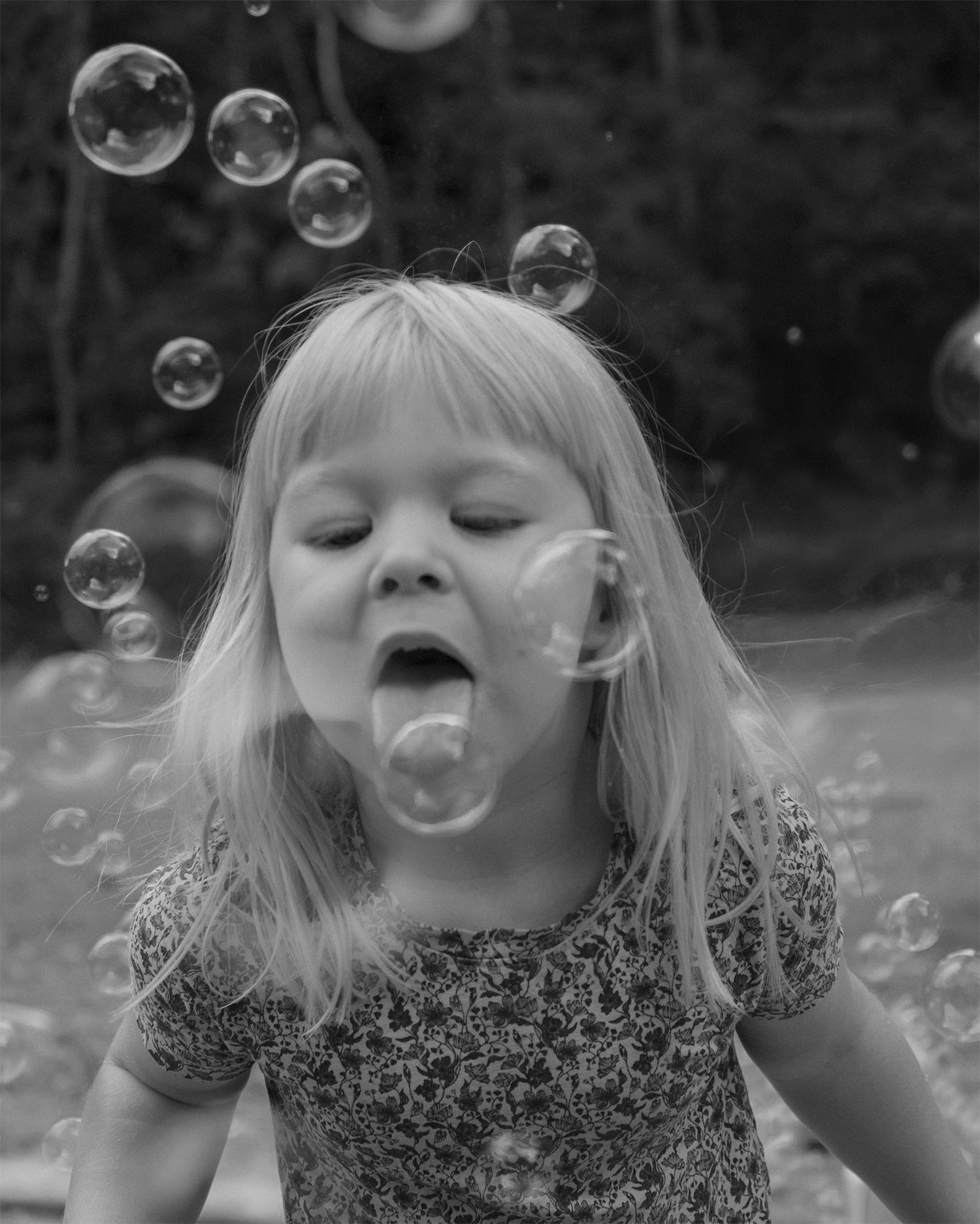 Black and white photo of a young blonde girl catching soap bubbles on her tongue outdoors.