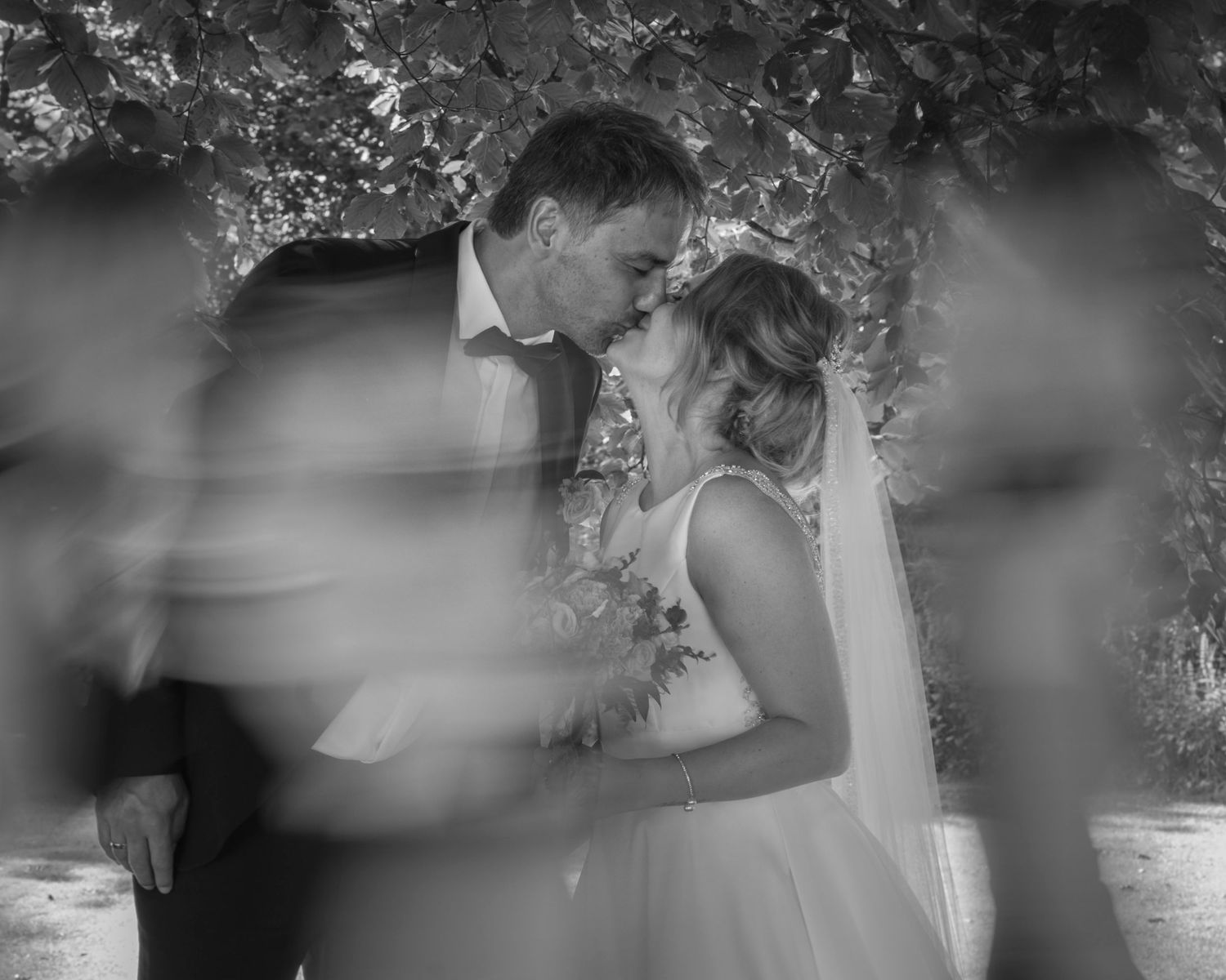 Black and white wedding photo of bride and groom sharing a kiss surrounded by blurred guests outdoors.
