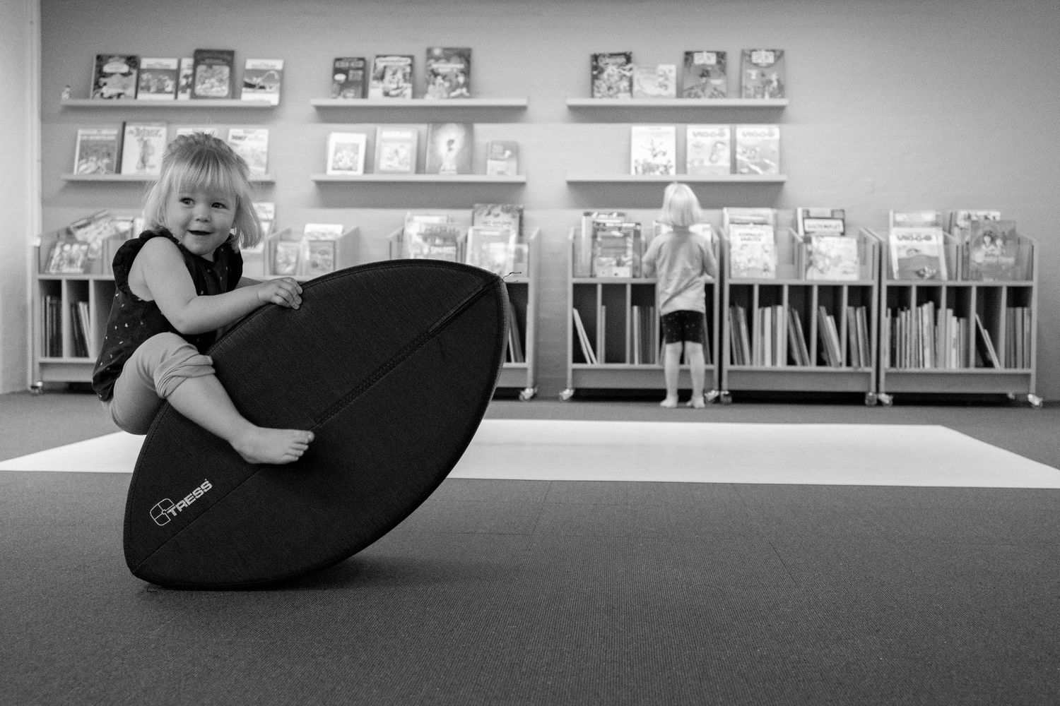 Young toddler playing on a black balance board in a library with bookshelves and a child browsing books in background.