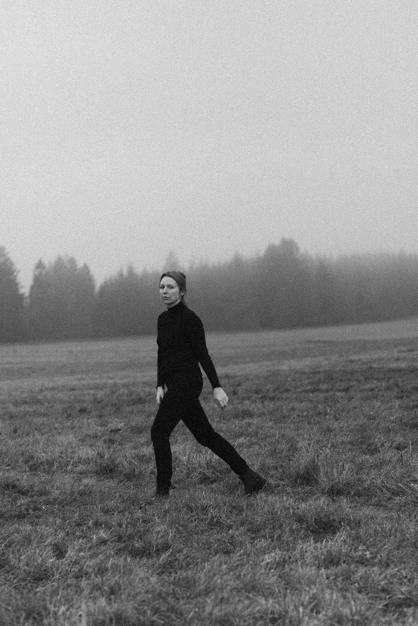 A person in all black walks through a misty, foggy field with trees in the background, black and white photo.