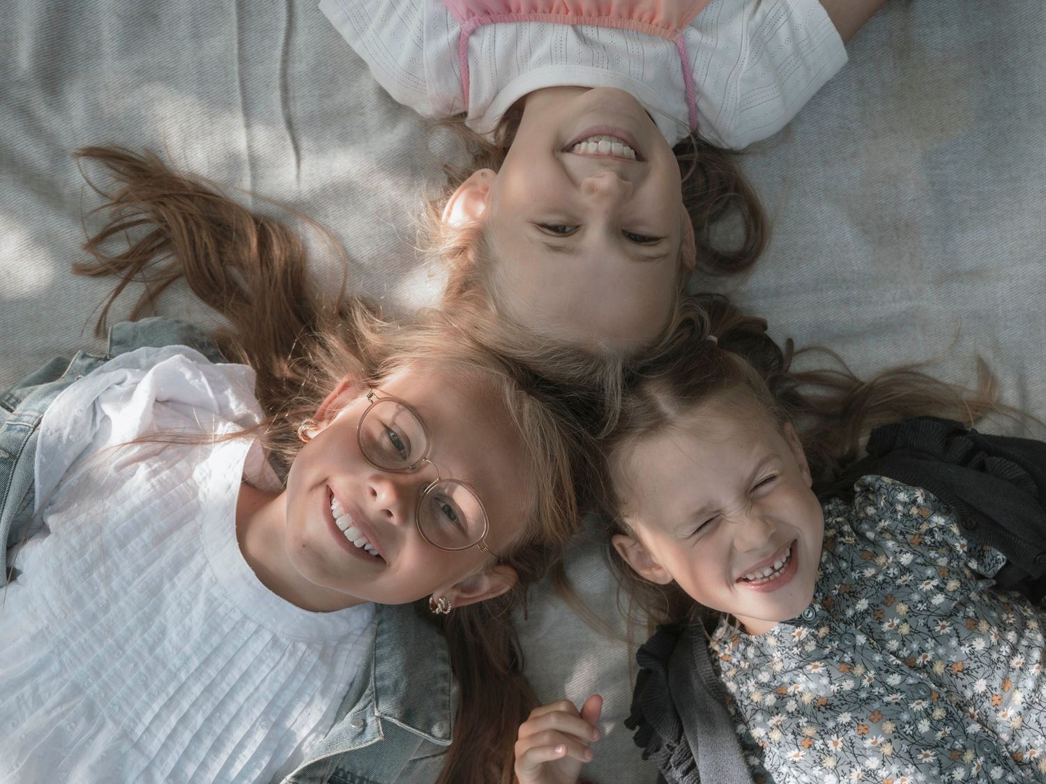 Three smiling girls lying on the ground with heads together, looking up at the camera in a joyful moment.