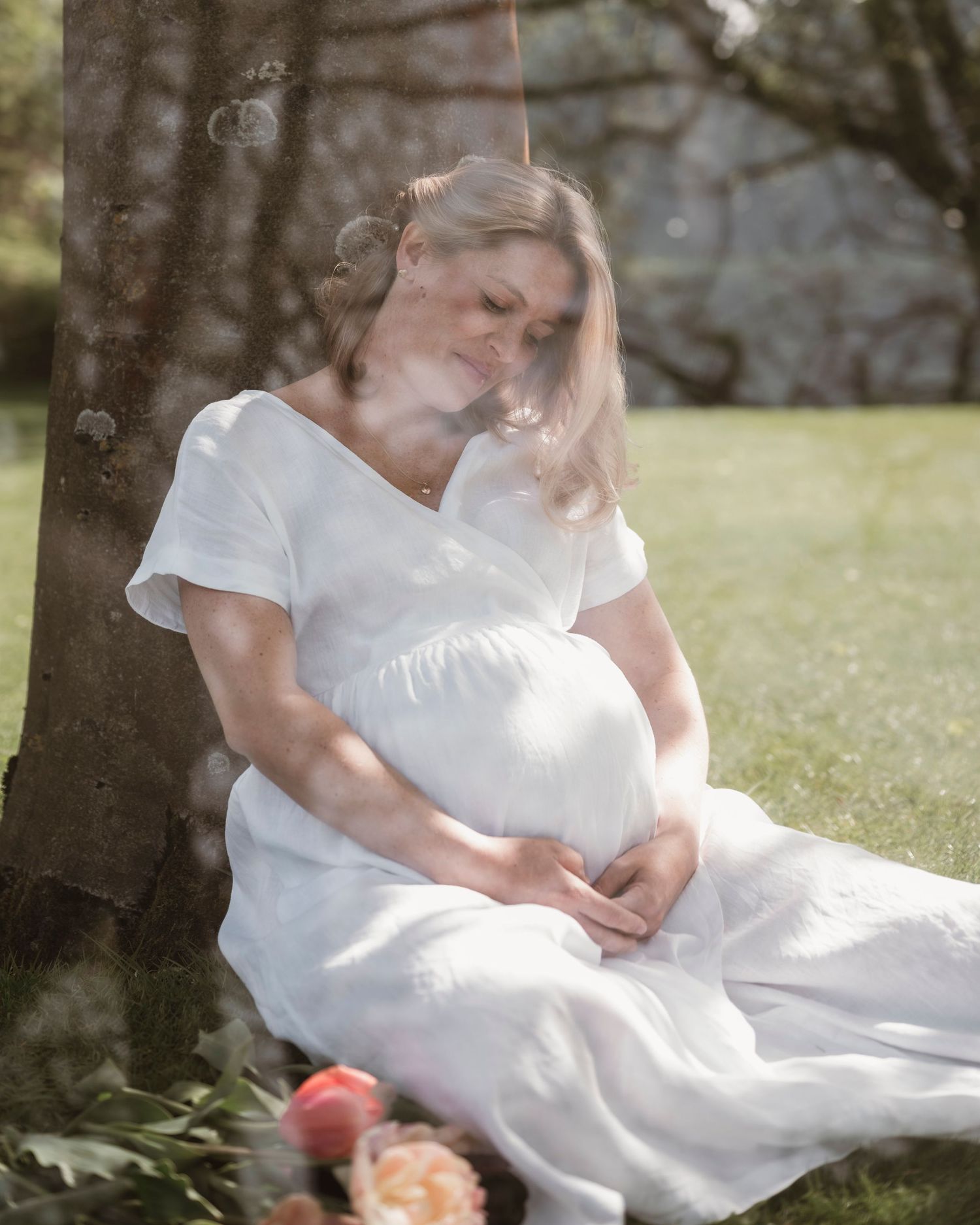Pregnant woman in white dress sitting against tree, cradling baby bump in a dreamy outdoor maternity photo.