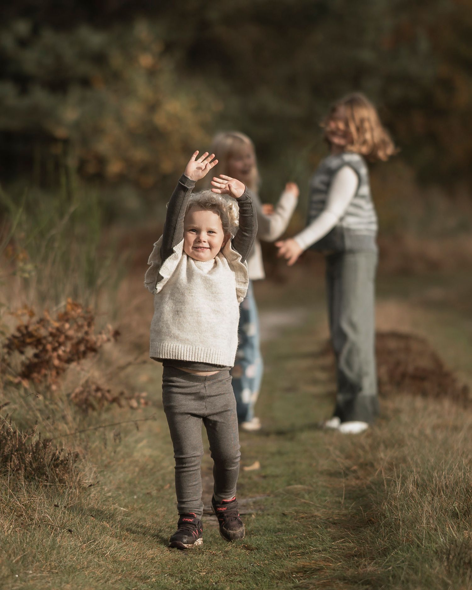 Three children playing outdoors on a nature path in autumn, with a smiling girl in foreground raising her arms joyfully.