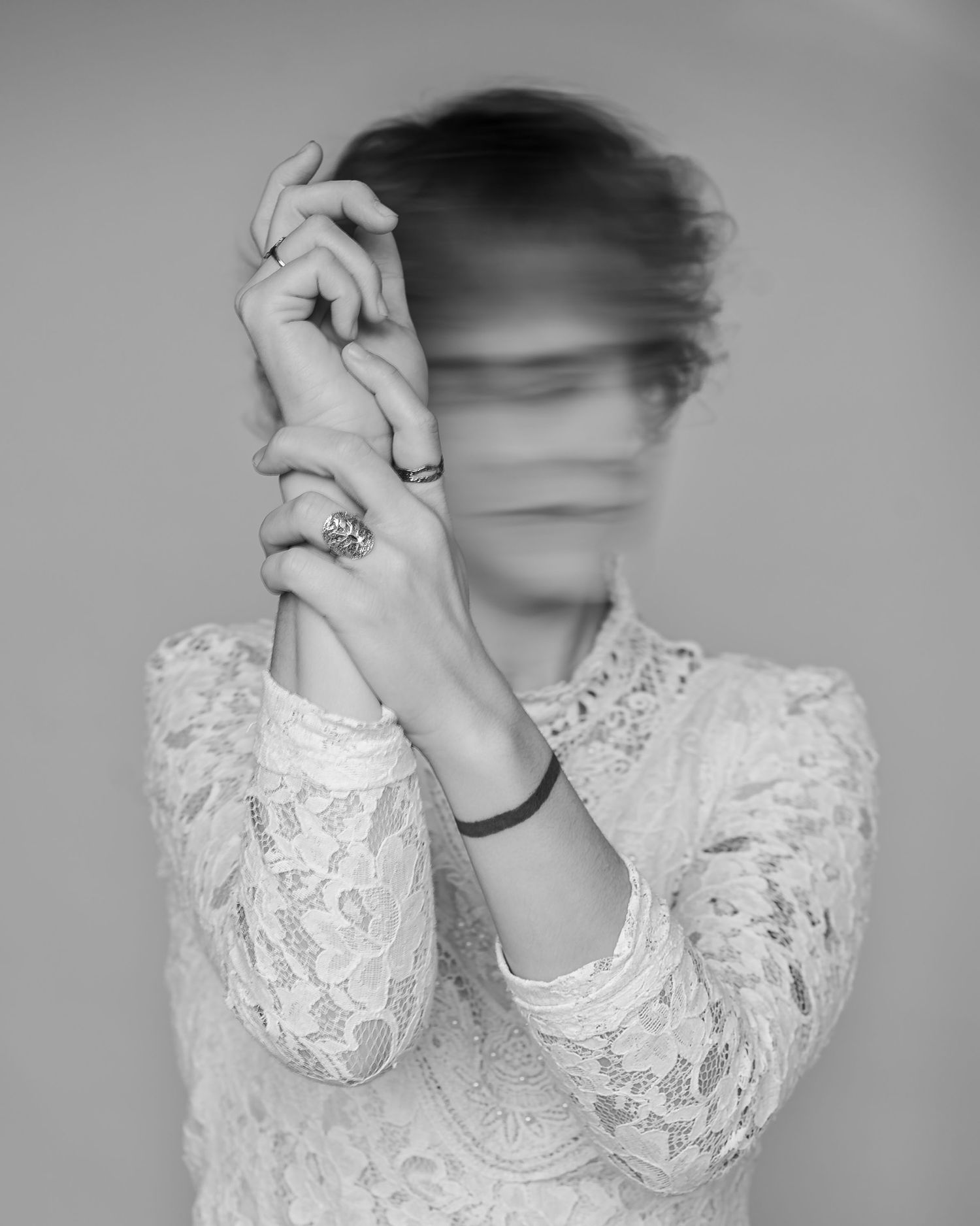 Black and white photo of a woman in lace dress with hands raised, face blurred by motion, wearing rings and bracelet.