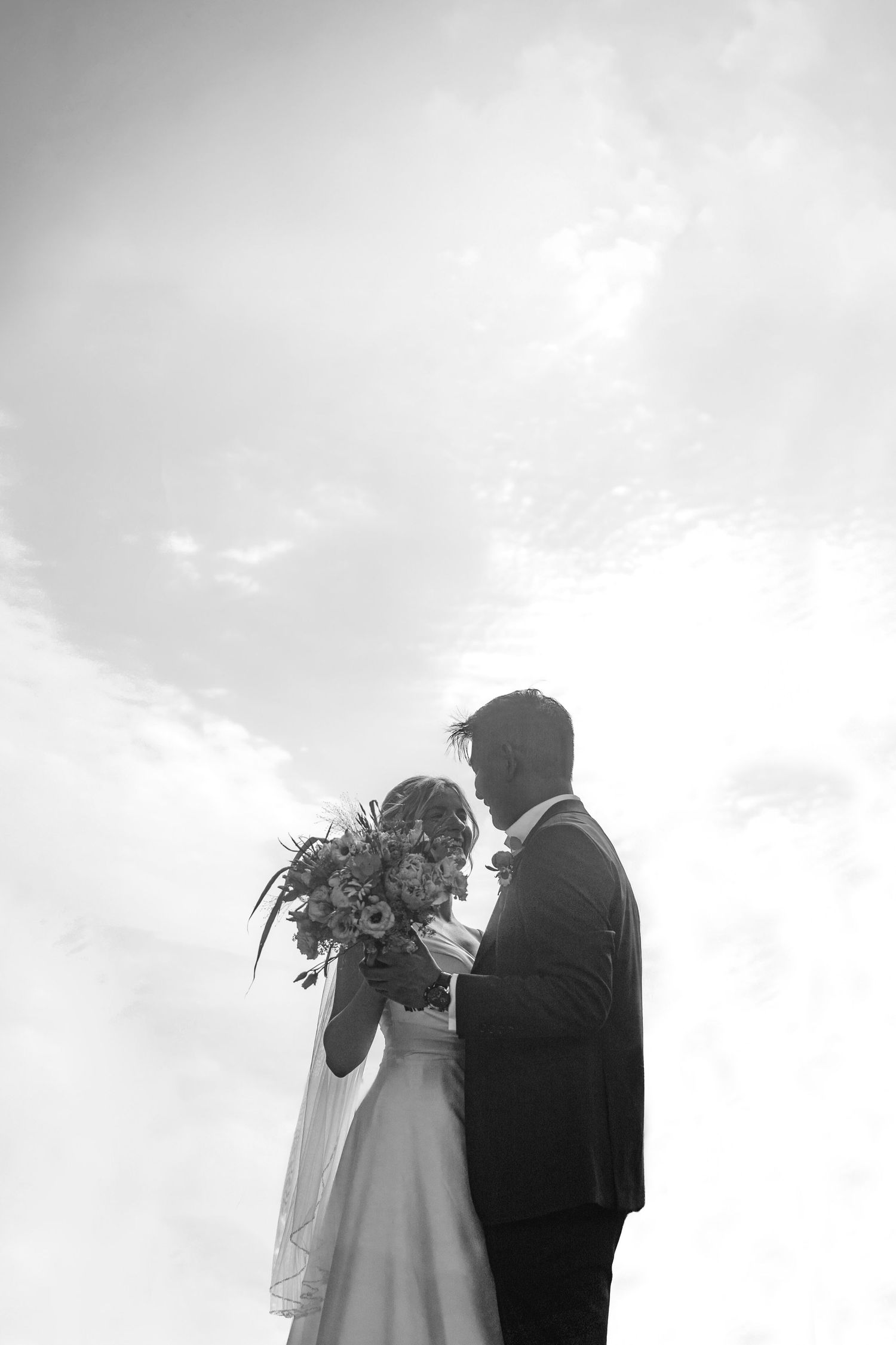 Black and white photo of bride and groom silhouetted against cloudy sky, bride holding floral bouquet.