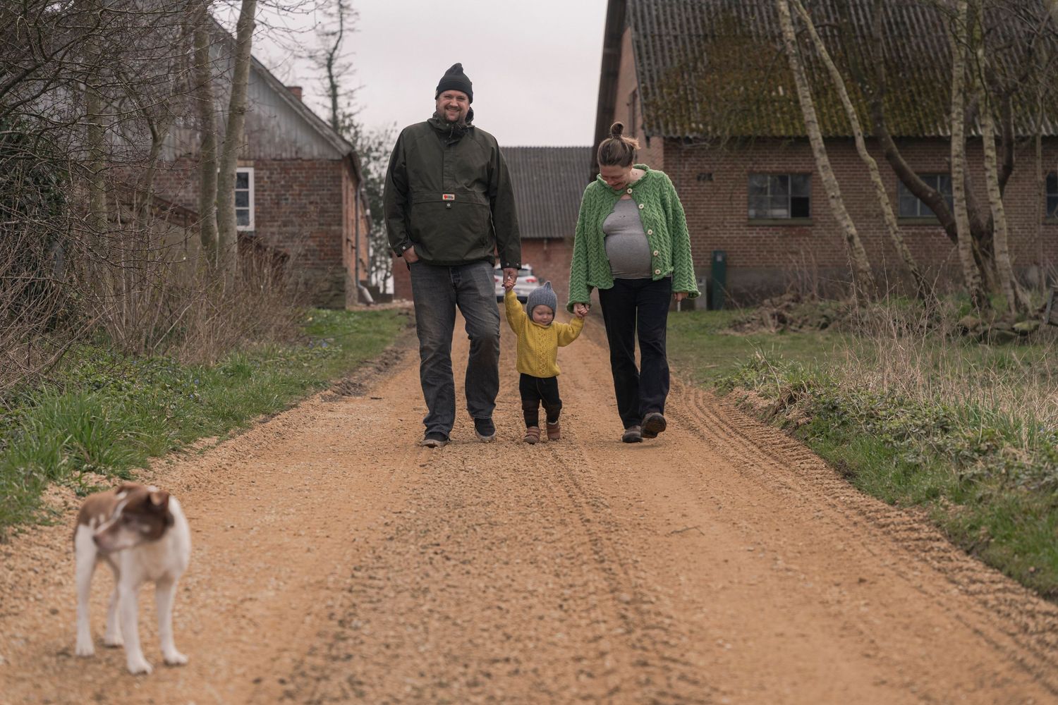 Family with toddler and dog walking on a rural dirt road past a farmhouse on a cloudy day.