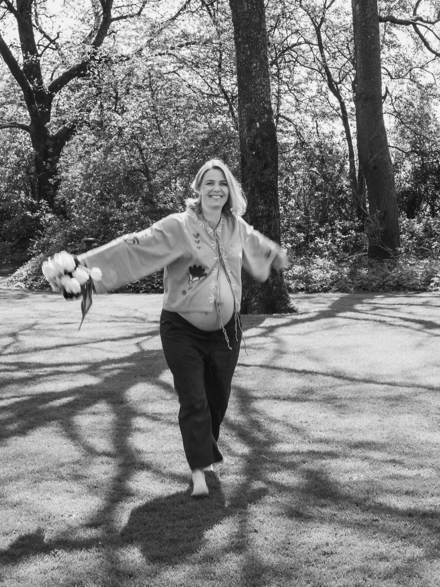 Black and white photo of a smiling pregnant woman dancing barefoot outdoors among trees.