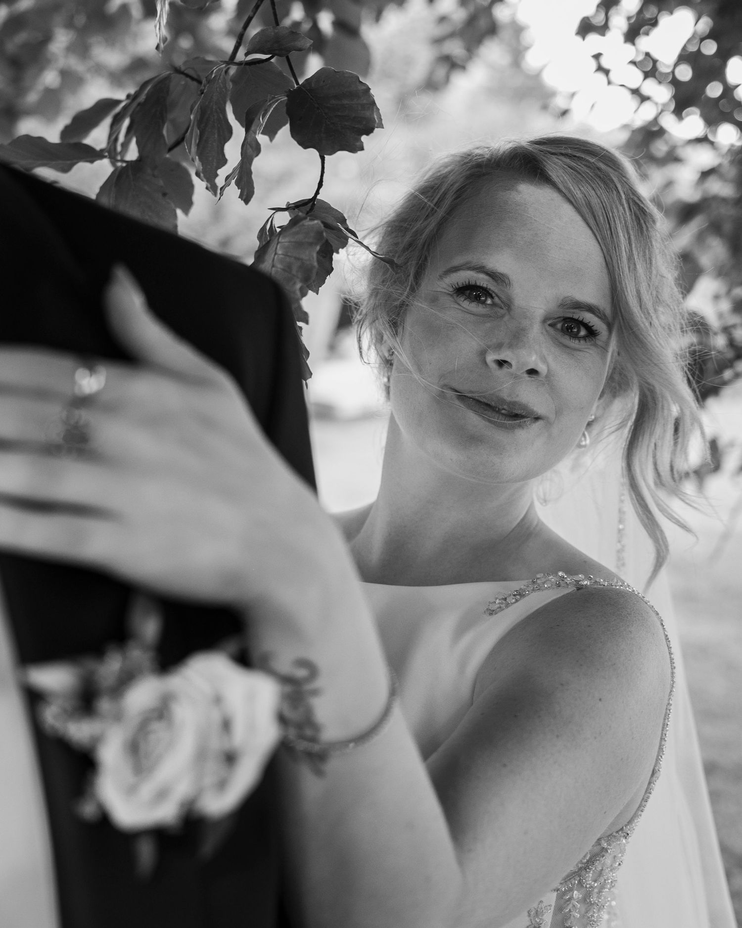 Black and white wedding photo of a smiling bride in a gown holding her groom, wearing a bracelet and earrings outdoors.