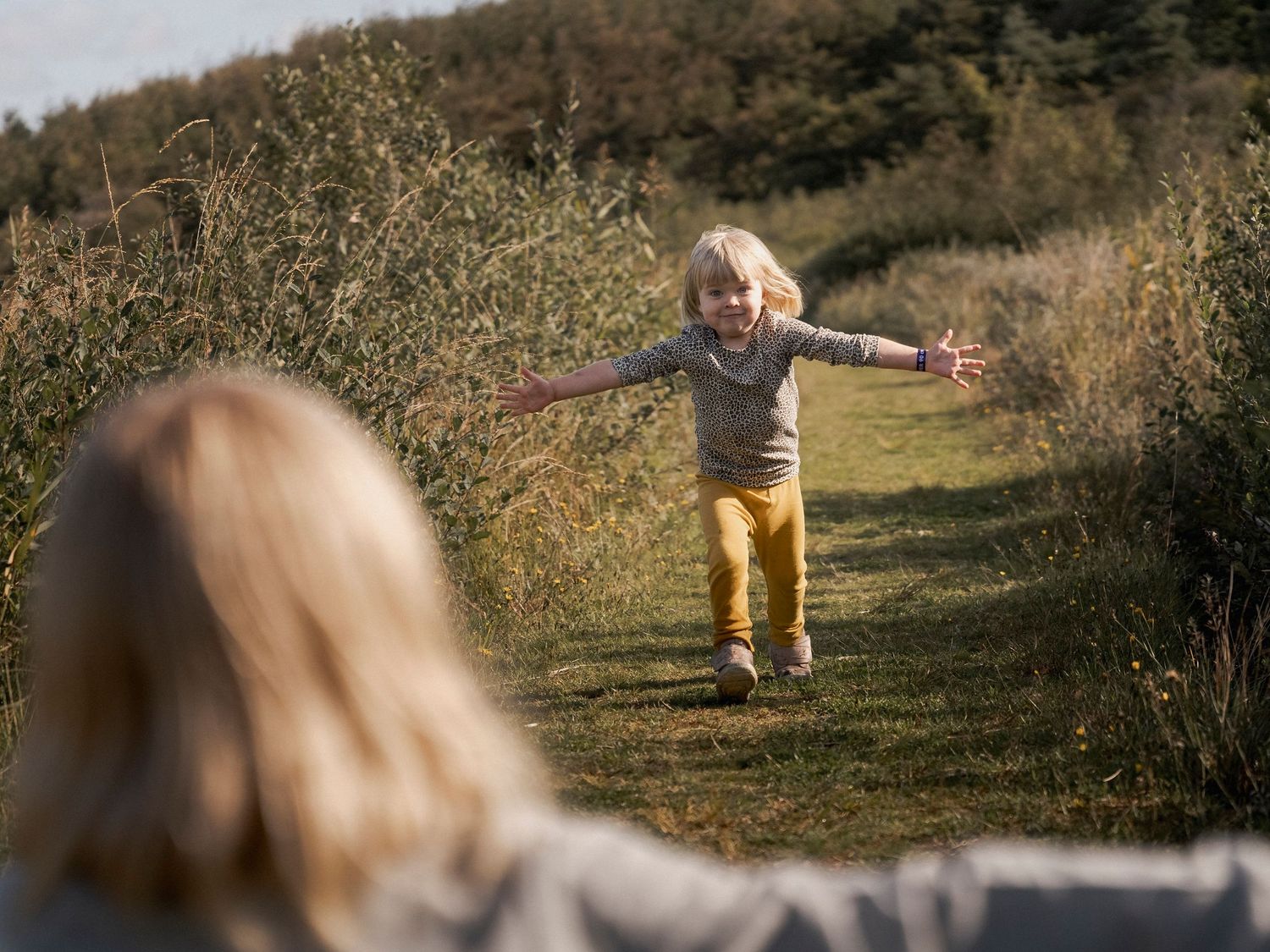 Joyful blonde child running with arms outstretched toward another child on a sunny green nature path.