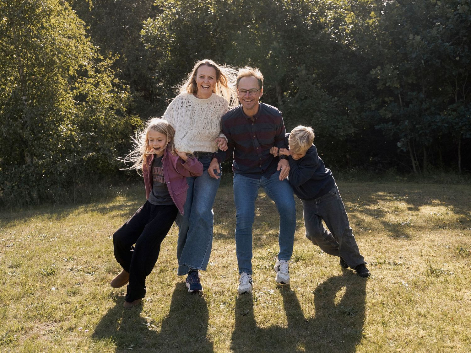 Family of four laughing and posing together outdoors on green grass with trees in the background on a sunny day.