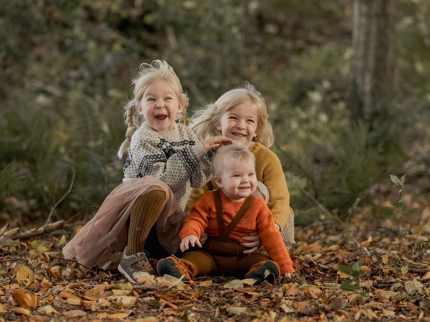 Three young children sitting in autumn leaves outdoors, smiling and laughing together in a fall forest setting.