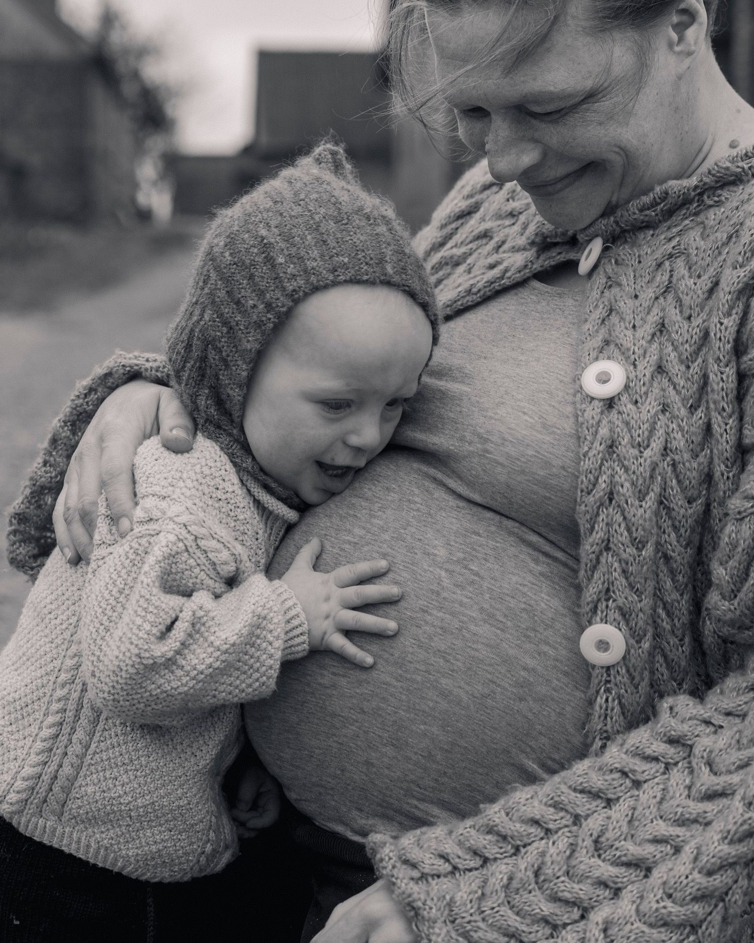 Black and white photo of a toddler hugging a pregnant woman's belly outdoors, both wearing knit sweaters.