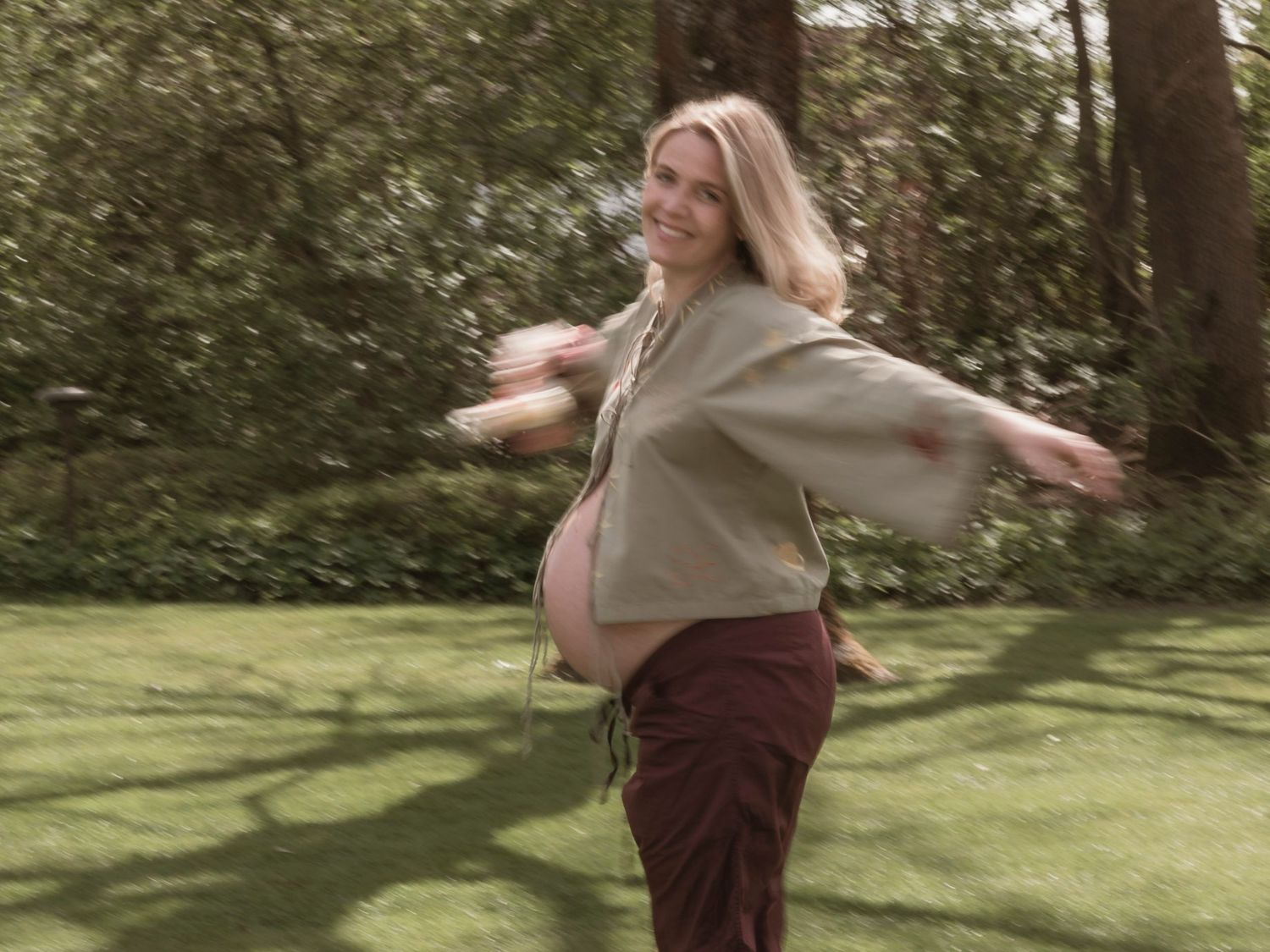 Smiling pregnant woman in sage top and burgundy skirt spinning outdoors in lush green garden setting.