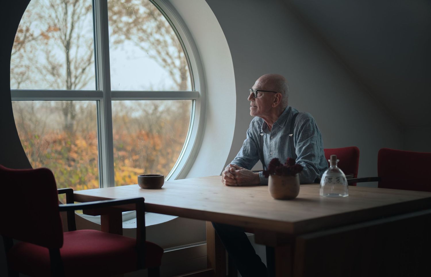 Elderly bald man with glasses sitting alone at a wooden table by a round window, gazing outside thoughtfully.